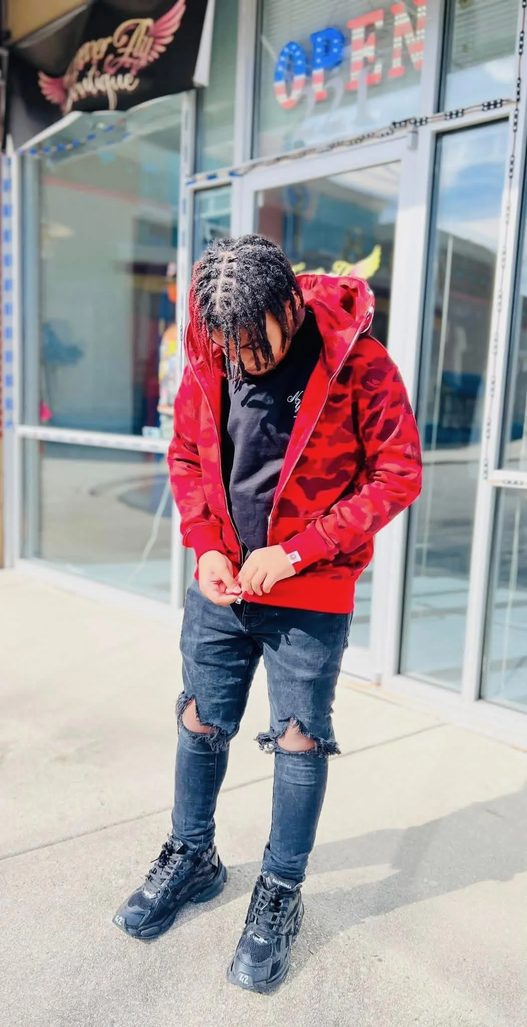 A young man with dreadlocks wearing a red camo hoodie, black t-shirt, ripped jeans, and black sneakers standing outside a storefront with glass doors and windows.