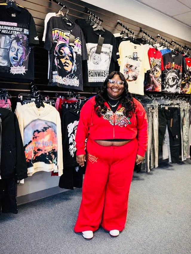 A smiling woman with glasses and long dark hair, dressed in a red tracksuit with a tiger design on the jacket, standing in a clothing store among various graphic T-shirts and hoodies displayed on the wall behind her.