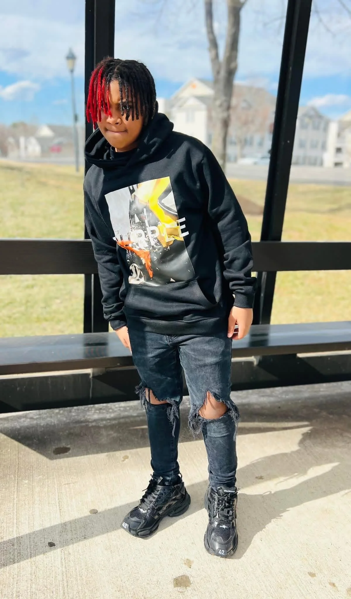 A young person with dreadlocks and red tips, wearing a black hoodie with a graphic on the front, ripped black jeans, and black sneakers, standing outdoors near a bench in a park or residential area on a sunny day.