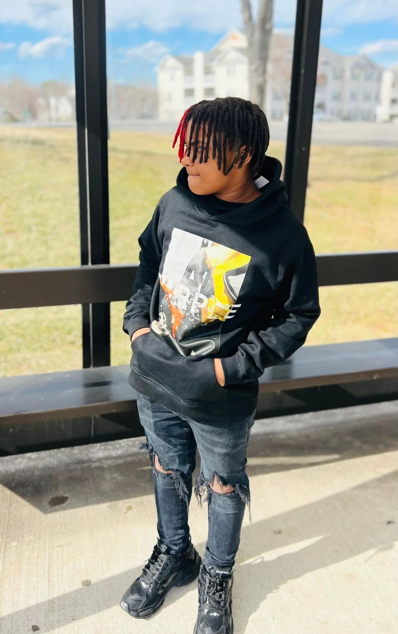 A young boy with dreadlocks and red tips, wearing a black hoodie with a graphic print, ripped jeans, and black sneakers, stands in a bus shelter with hands in pockets, looking to the side with a contemplative expression, outdoors on a sunny day.