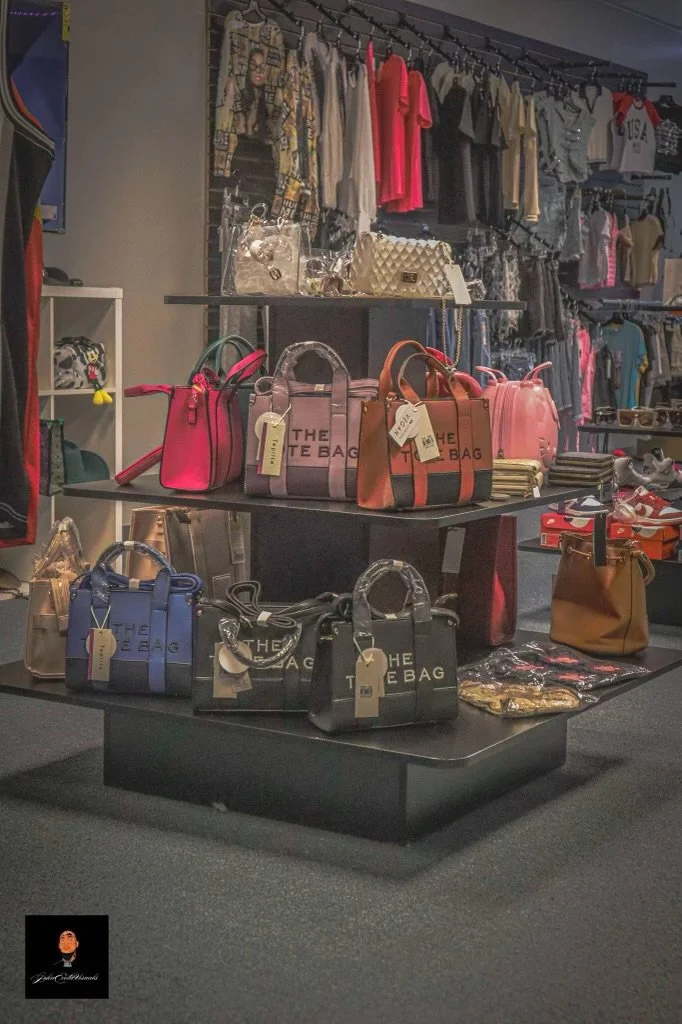 Display of colorful handbags on a black retail table in a clothing store, with clothes hanging on racks in the background.