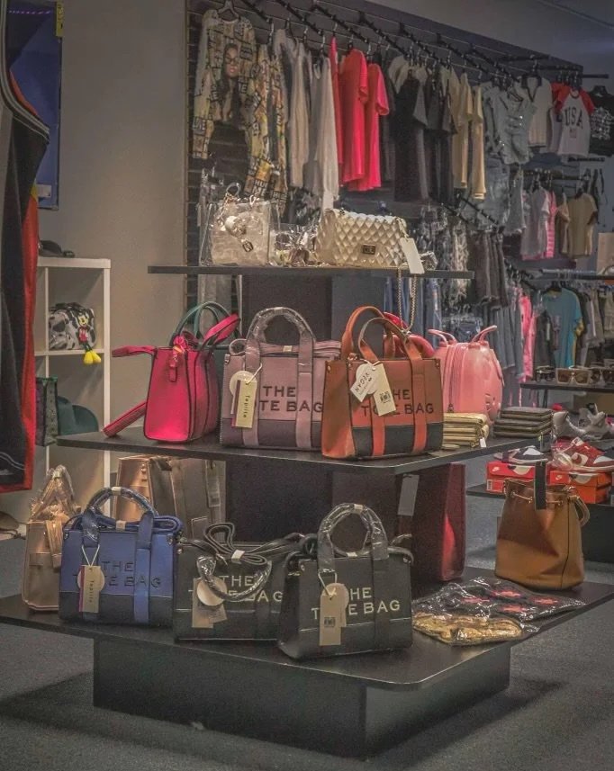 Display of colorful handbags on a two-tiered table inside a retail store, with clothing and accessories in the background.