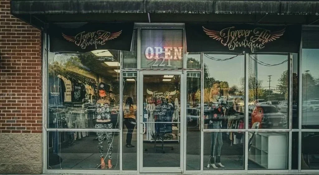 storefront of Forever Fly Boutique with clothing inside and an illuminated 'OPEN' sign in the window.