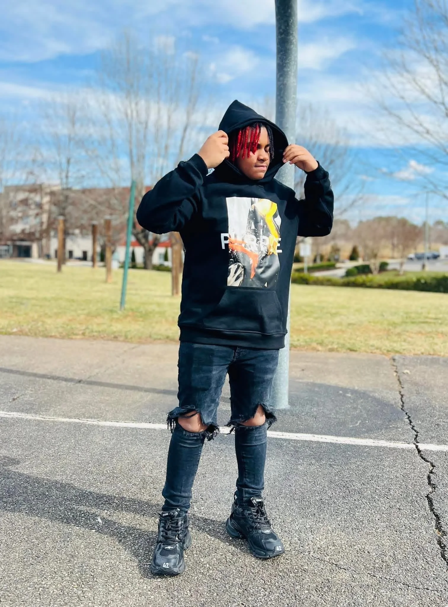 A young man with red-tinted hair in a hoodie, standing outdoors on a cloudy day, adjusting his hoodie over his head. He is wearing a black hoodie with a graphic print, ripped jeans, and sneakers.