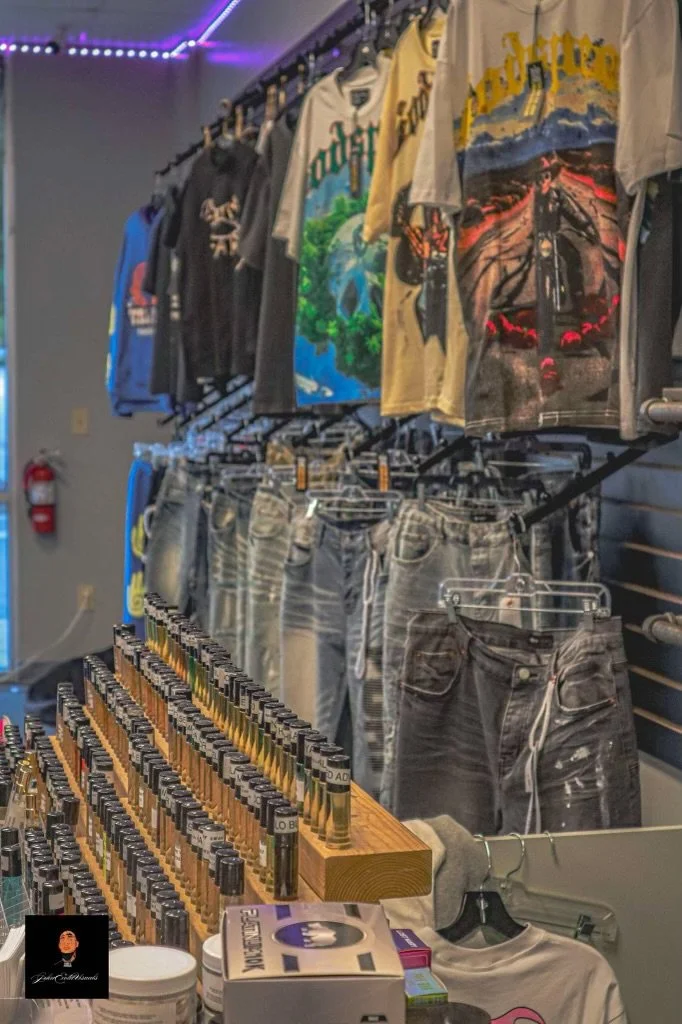 Display of graphic T-shirts with various artwork, including landscapes and racing themes, paired with distressed denim jeans on hangers, and a display of small bottles and wellness products on a wooden rack in a retail store.