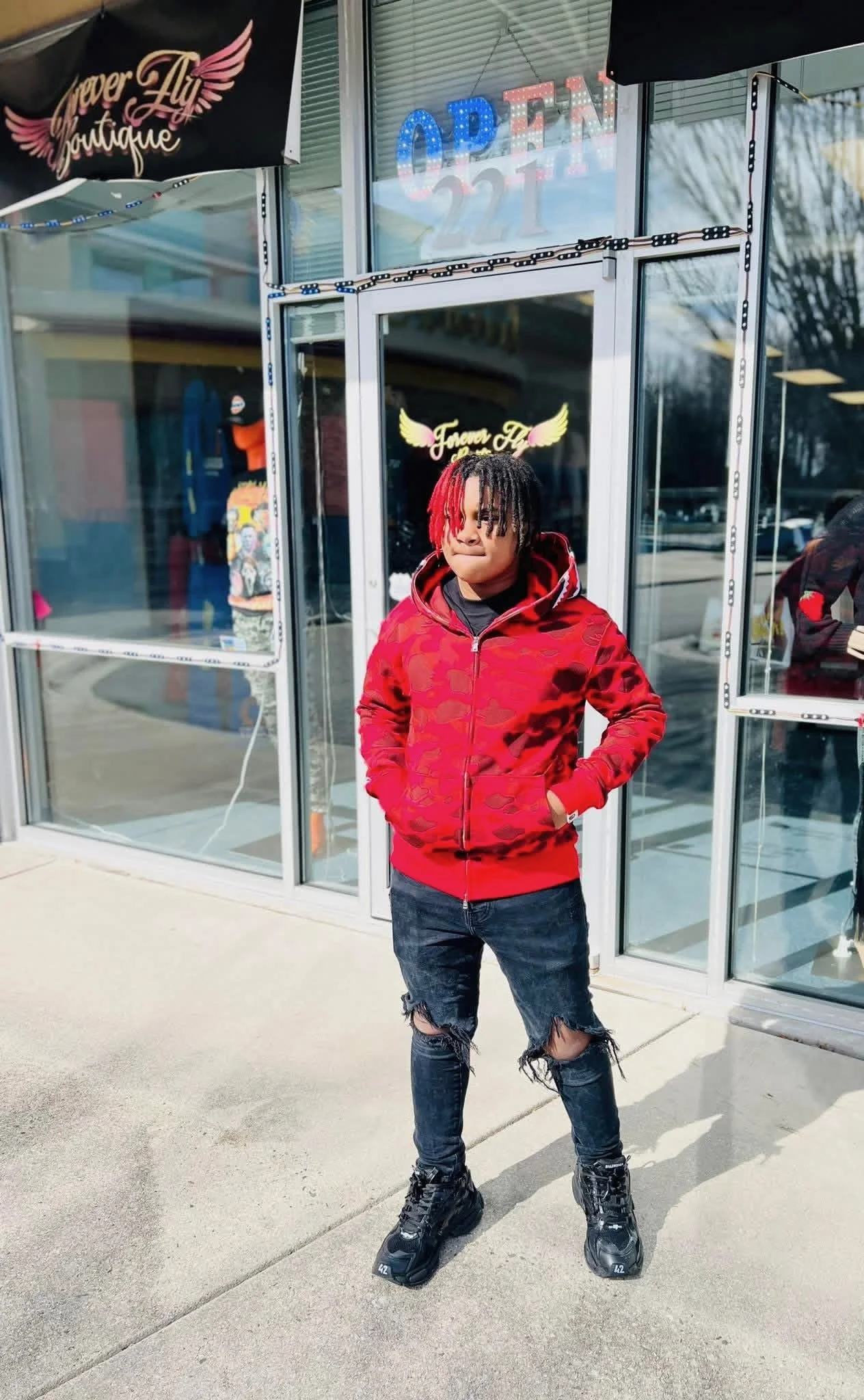Young person with red and black dreadlocks standing outside a boutique store wearing a red camouflage hoodie, ripped jeans, and black sneakers, with store signage and glass windows behind.
