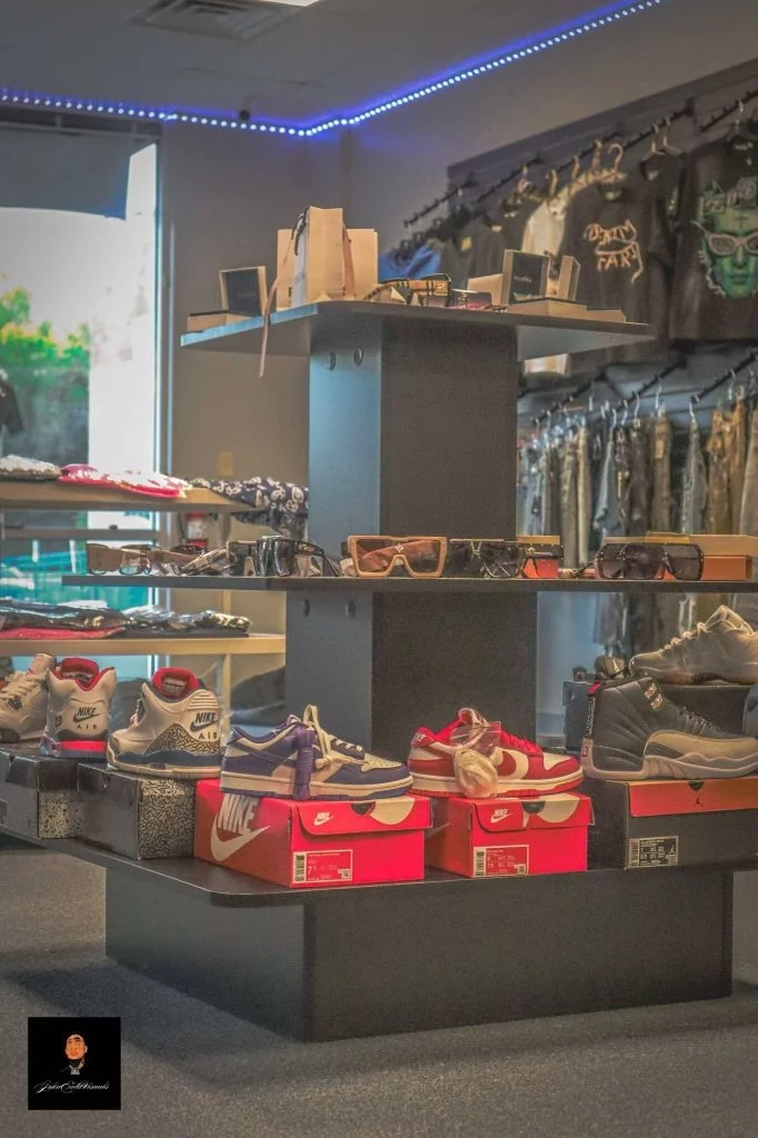 Display of sneakers, sunglasses, and accessories in a retail store with clothing in the background.