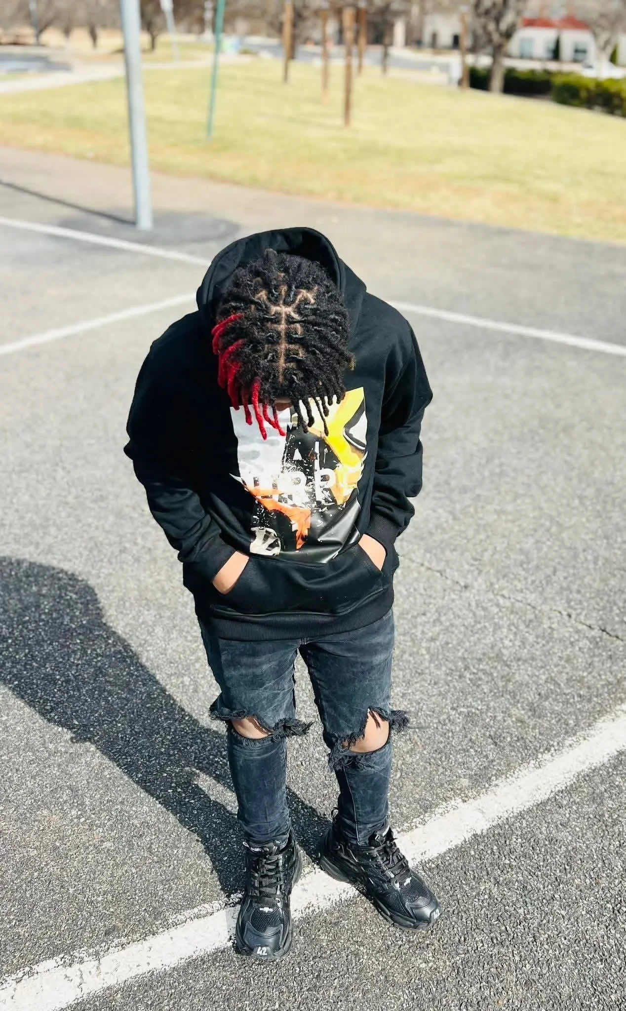Young person with twisted hair, wearing a black hoodie, ripped jeans, and black sneakers, standing with hands in pockets on a paved outdoor area with a grassy background and trees.