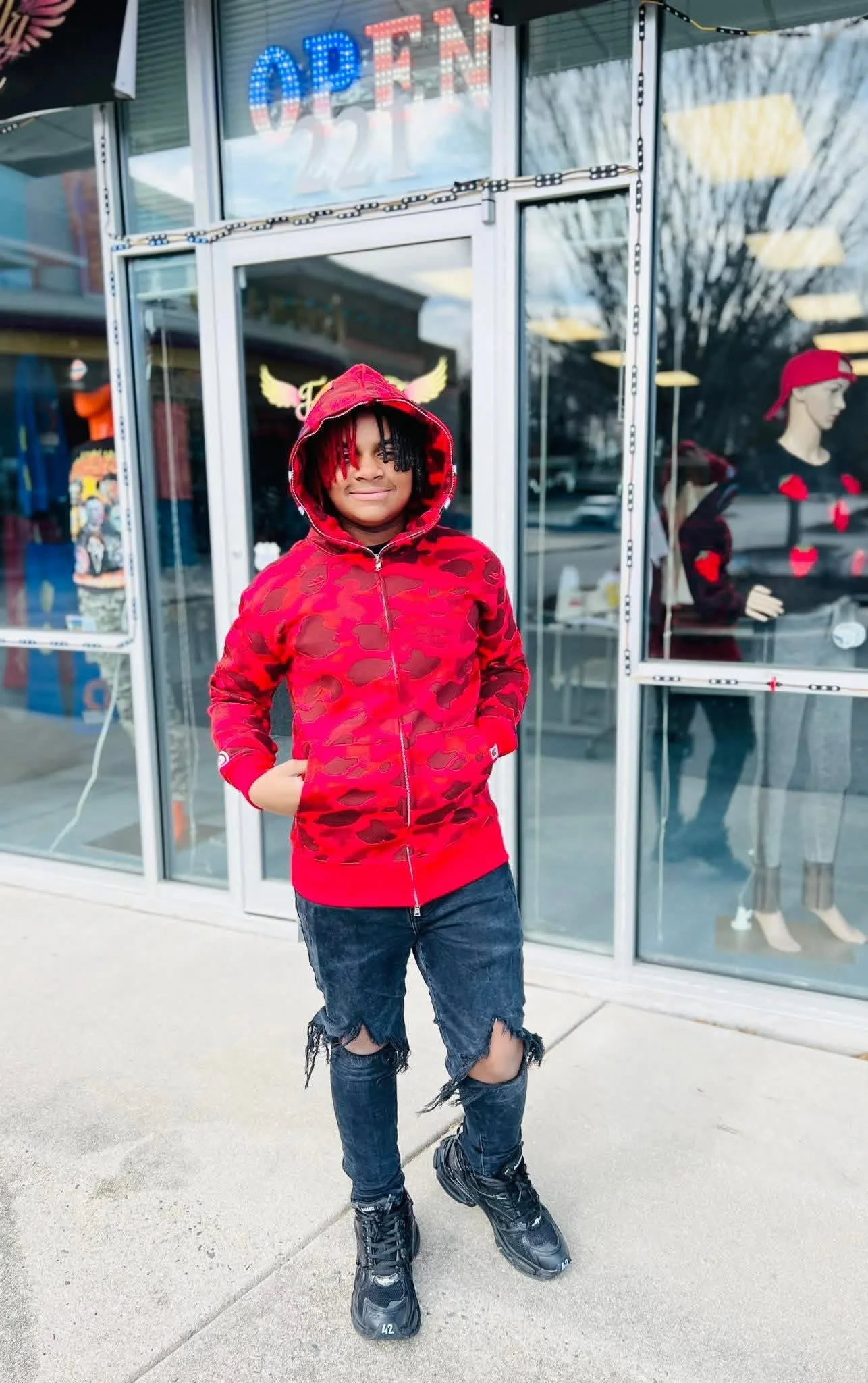 A young person standing outdoors in front of a store window with an 'OPEN' neon sign. They are wearing a red camo hoodie with a hood, ripped jeans, and black sneakers, smiling slightly with their hands in their hoodie pockets.