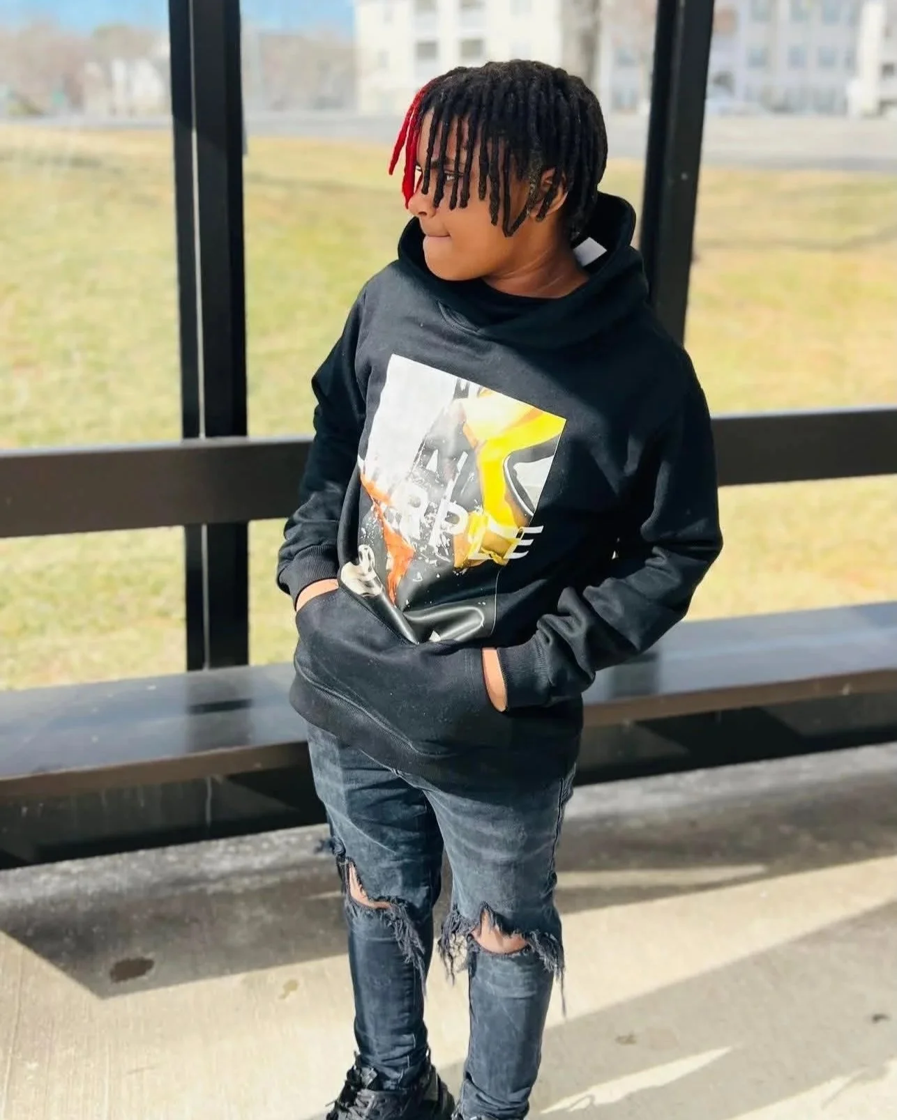 A young person with dreadlocks, with red tips, wearing a black hoodie with a graphic print, ripped jeans, and sneakers, standing outdoors near a black metal and glass shelter.