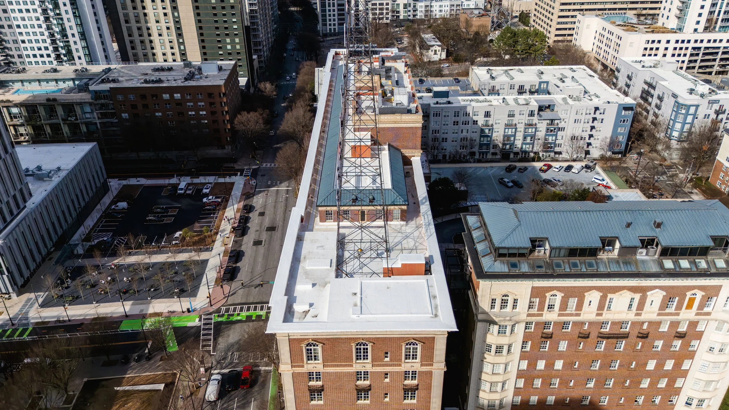 An aerial view of a cityscape showing multiple high-rise buildings, streets, parking lots, and a rooftop with a large antenna structure, during daytime.