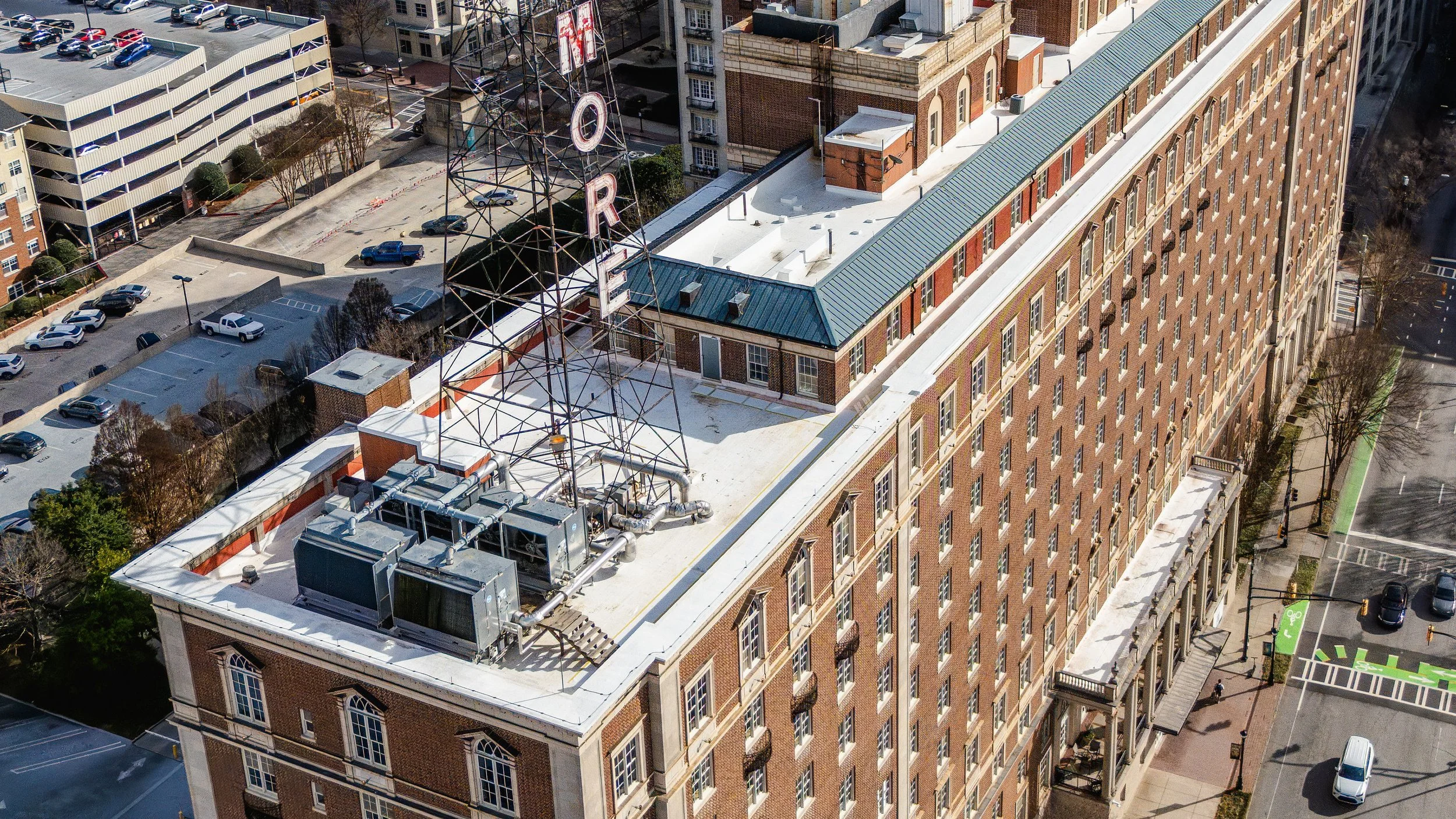 An aerial view of a tall brick building with a green metal roof, featuring a large metal sign structure for a theater's marquee that reads 'THE ORPHEUM.' The rooftop contains vents, ducts, and equipment. Adjacent to the building is a parking lot with parked cars and streets lined with trees.