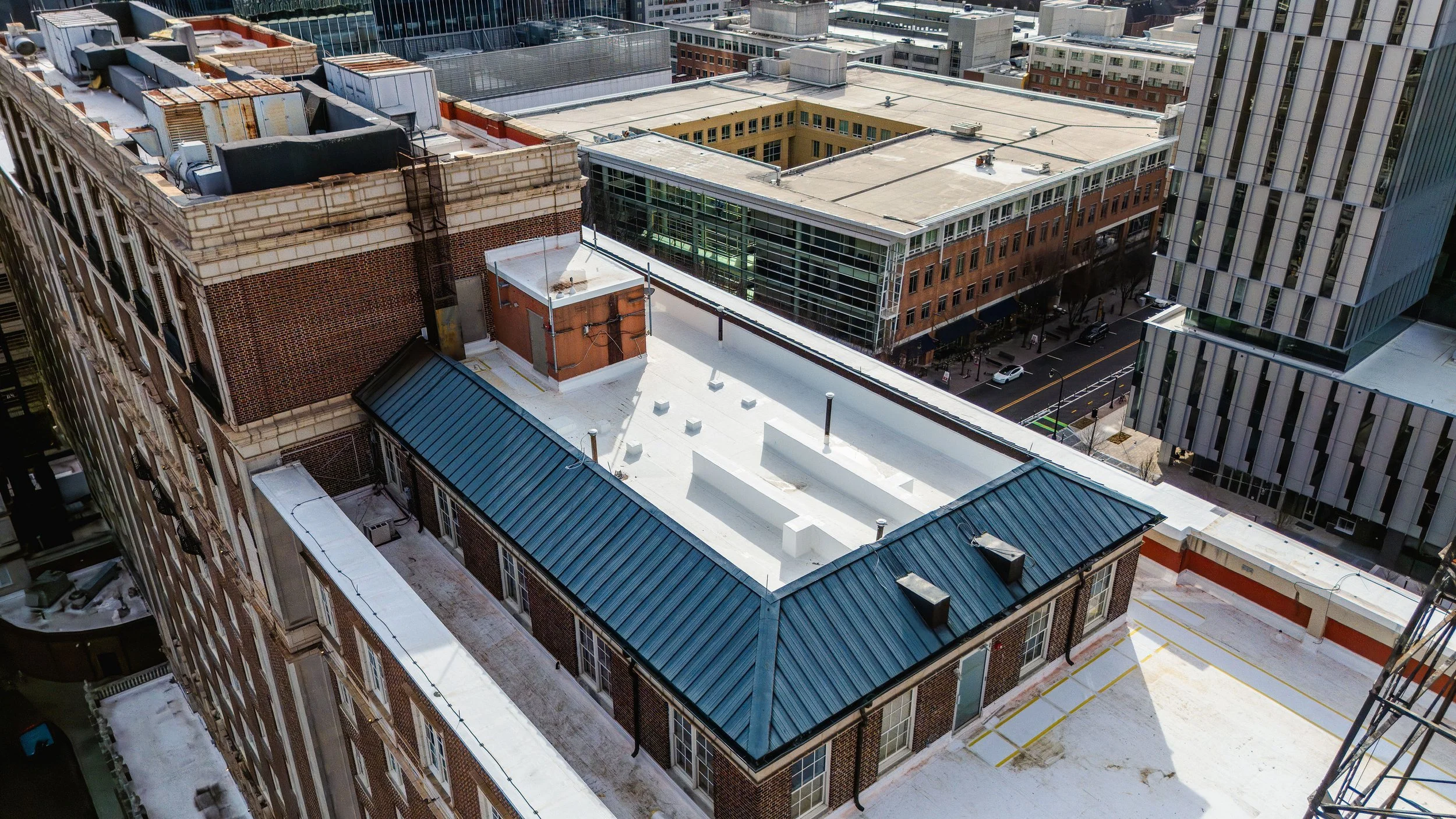 A cityscape viewed from above showing rooftops of various buildings, including a red brick building with a green roof, in an urban downtown area with taller modern office buildings in the background.