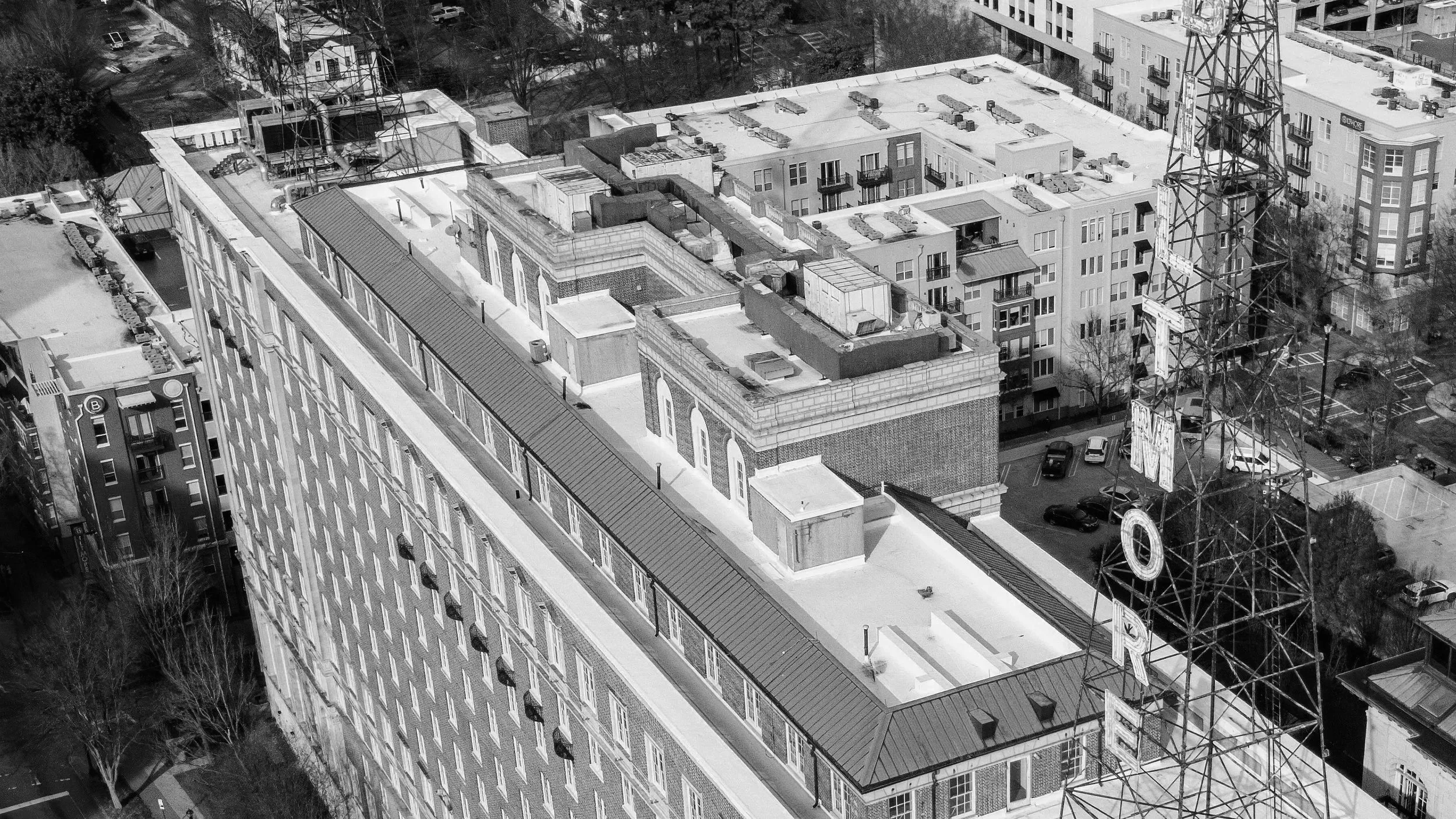 Black and white aerial view of a cityscape with a prominent large building topped with a water tank and multiple rooftop structures. A tall radio tower is nearby, with several parked cars and nearby buildings visible.