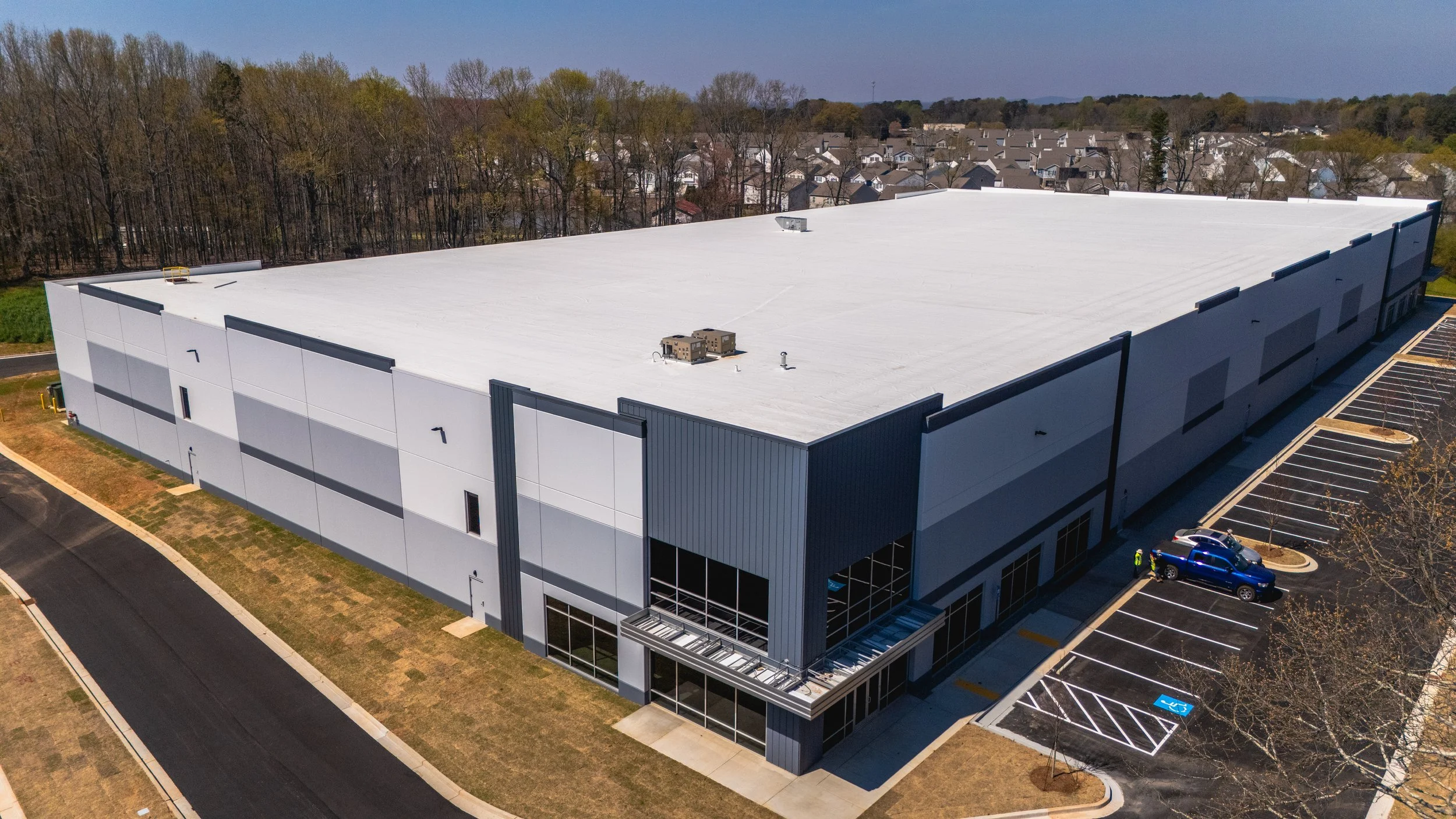 Aerial view of a large commercial building with a white flat roof, parking lot with multiple cars, and surrounded by trees and a neighborhood in the background.