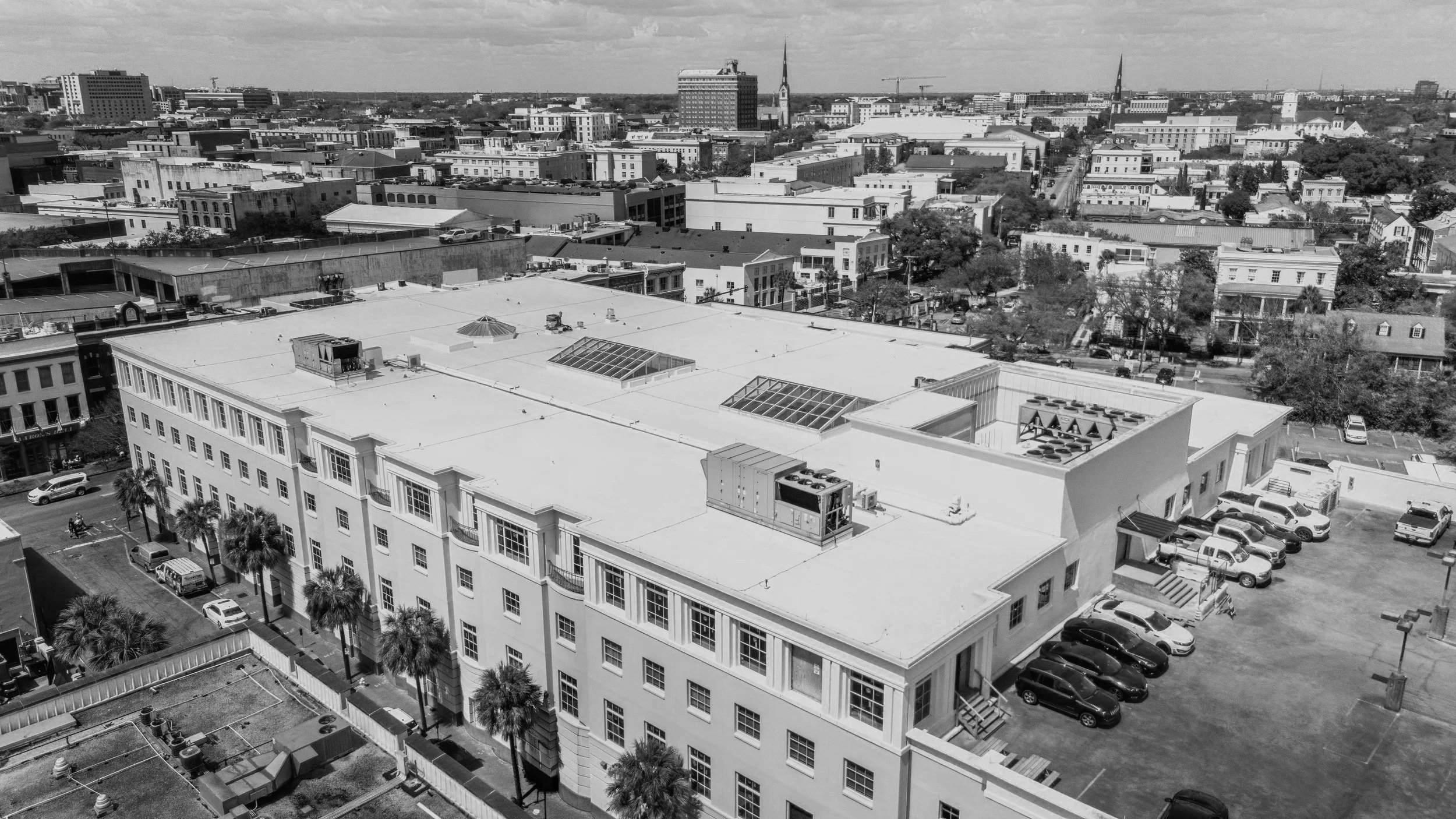 Black and white aerial view of cityscape with multiple buildings, parking lots, and trees. Tall buildings and church steeples in the background under cloudy sky.