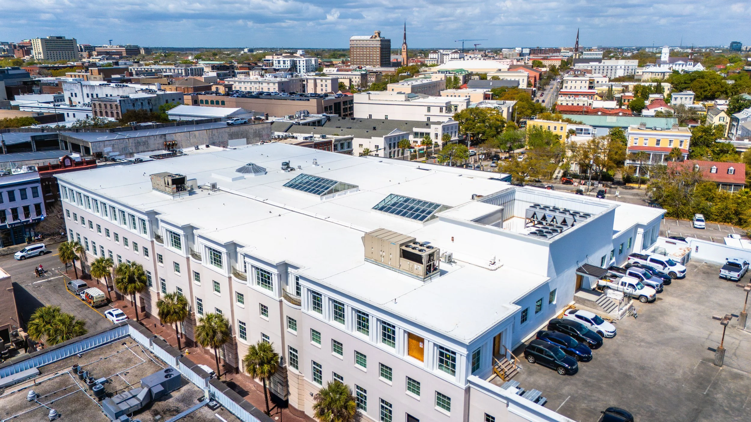 Aerial view of a cityscape with a white building in the foreground, featuring a flat roof with skylights and HVAC units, surrounded by parking lots and palm trees. The background shows various buildings, church steeples, and a partly cloudy sky.