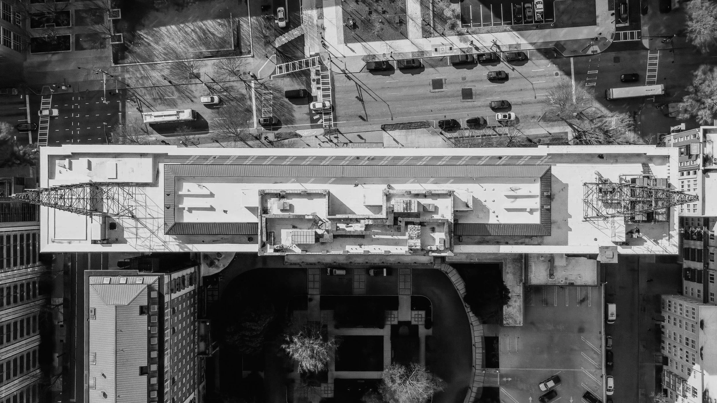 Aerial view of a city street with parked cars, sidewalks, and buildings, captured in black and white.