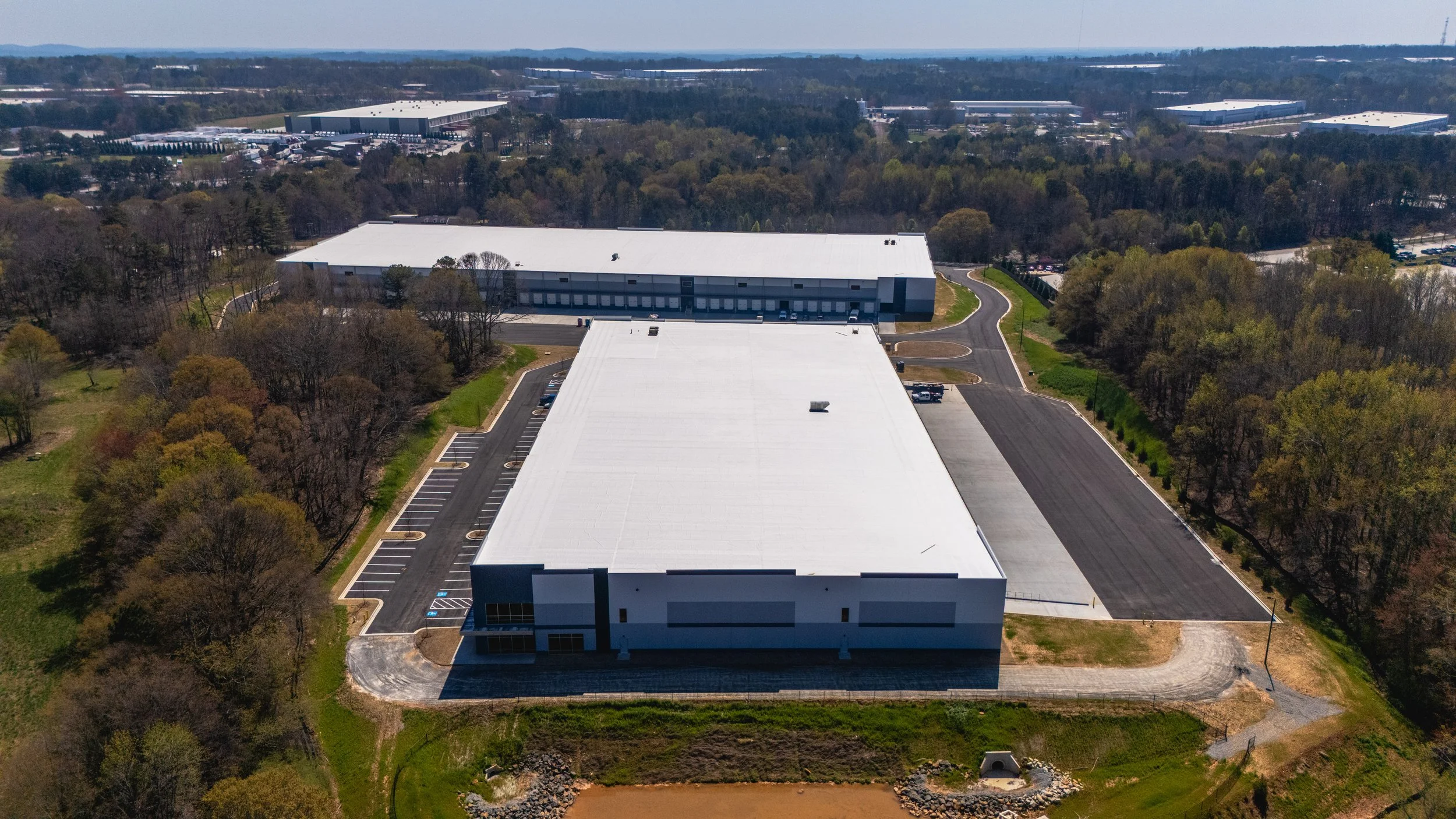 An aerial view of a large warehouse with a white roof surrounded by parking lots and trees, in a commercial area with other industrial buildings in the background.