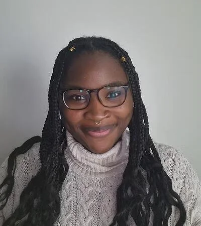 A woman with glasses and a septum ring, smiling in front of a plain light-colored wall, wearing a beige knitted turtleneck sweater and having long braided hair with small gold accessories.