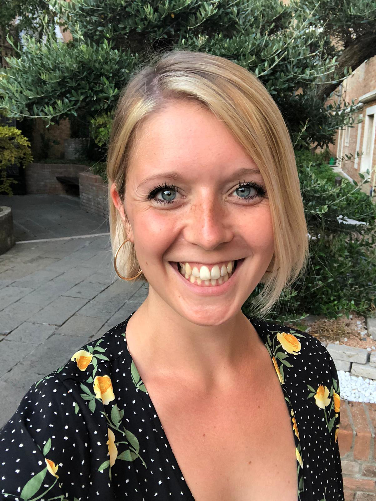 A smiling woman with short blonde hair, blue eyes, and pierced ears, wearing a black dress with yellow flowers and white polka dots, standing outdoors in front of green bushes and trees.