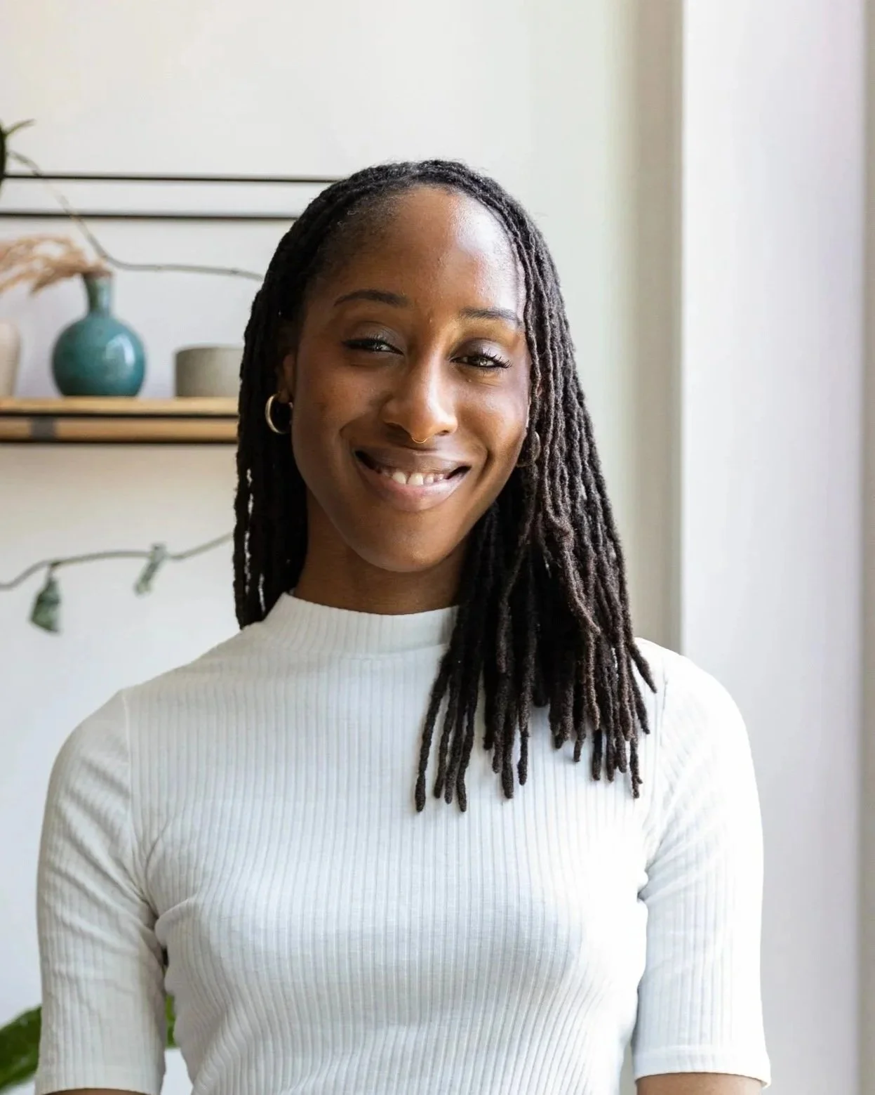 A woman with dark skin and long locs smiling in a room with a white wall and shelves holding vases and decorative items.
