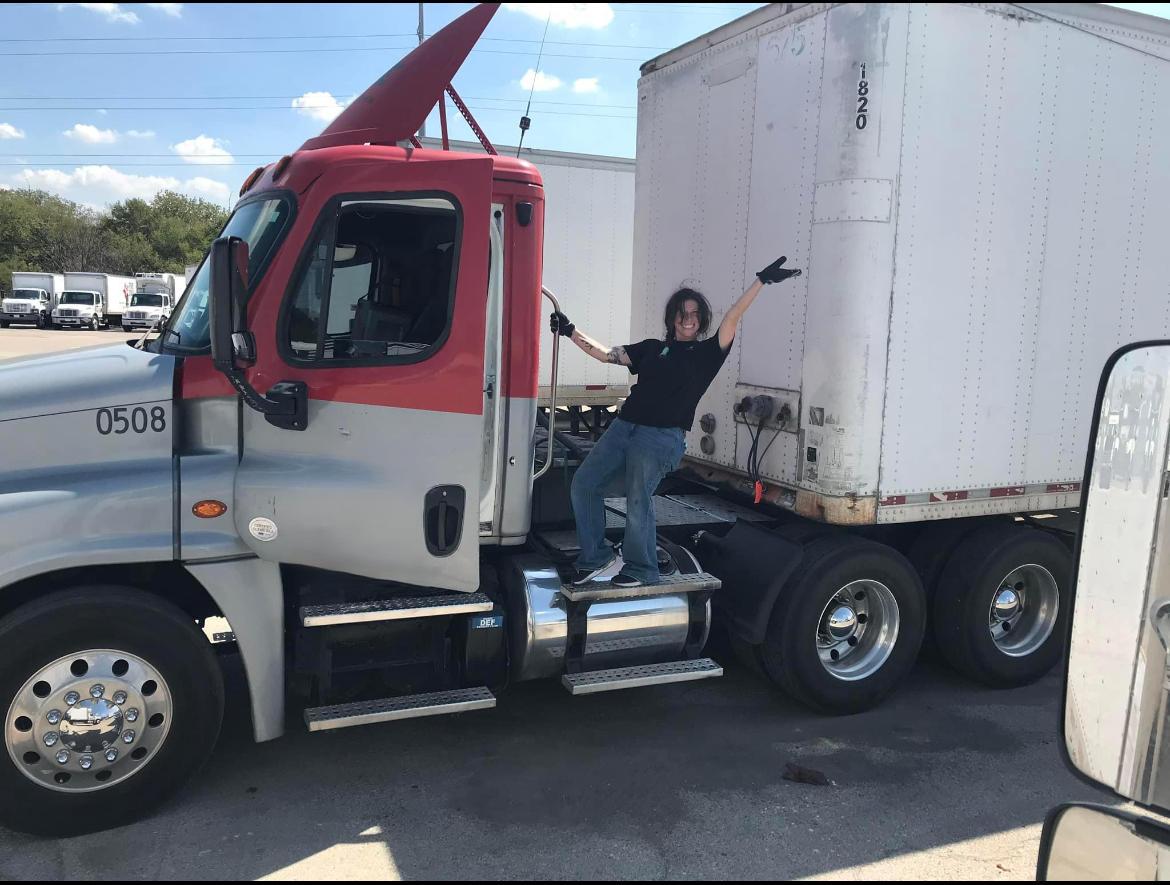 A woman standing on the side step of a semi-truck, smiling and pointing at the truck, with her arms raised in joy.