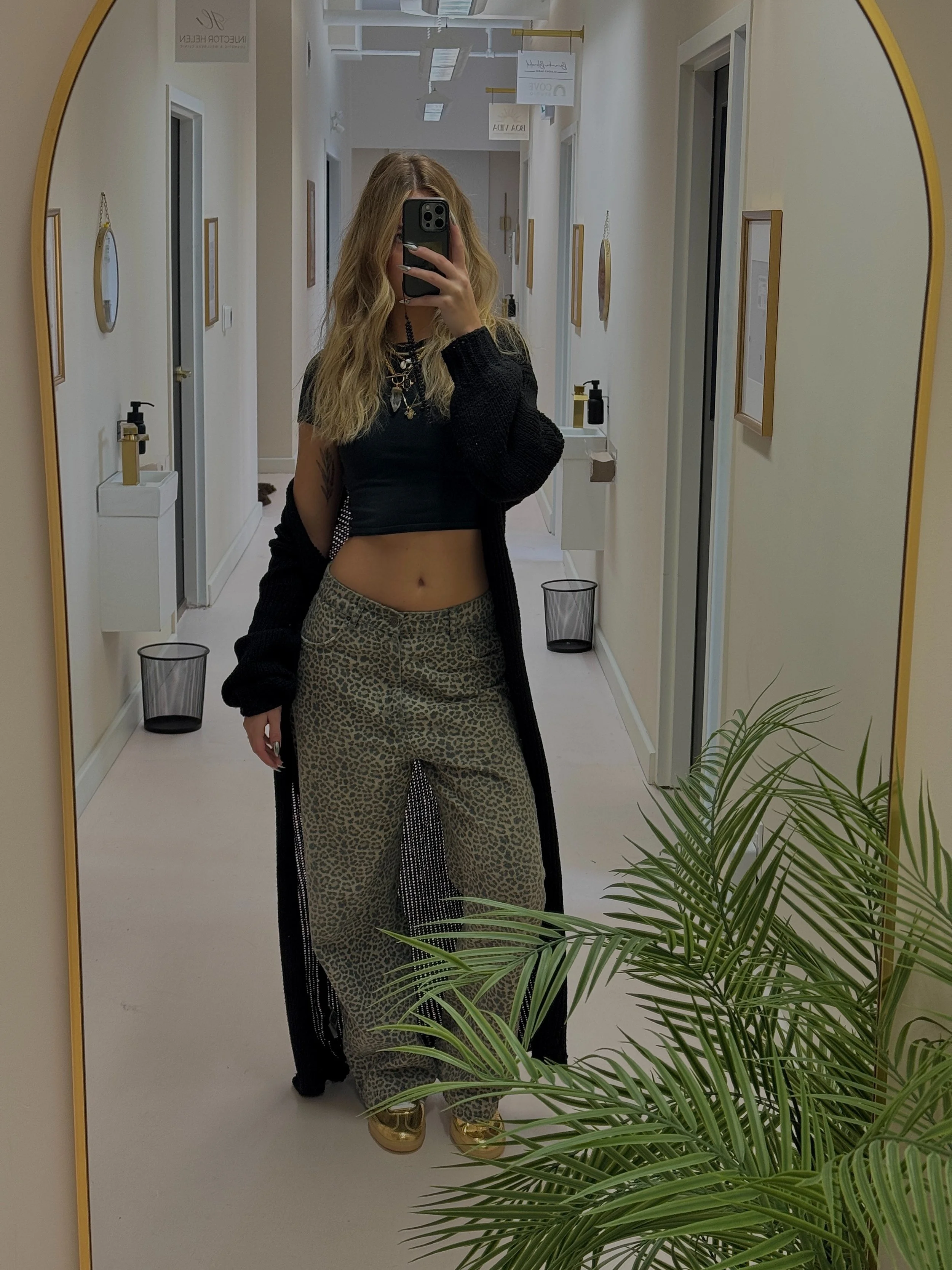 A woman with long wavy blonde hair taking a mirror selfie in a well-lit corridor. She is wearing a black crop top, leopard print pants, a long black cardigan, and gold shoes. A plant is visible in the foreground, and the corridor has framed pictures on the walls and small sinks with soap dispensers.