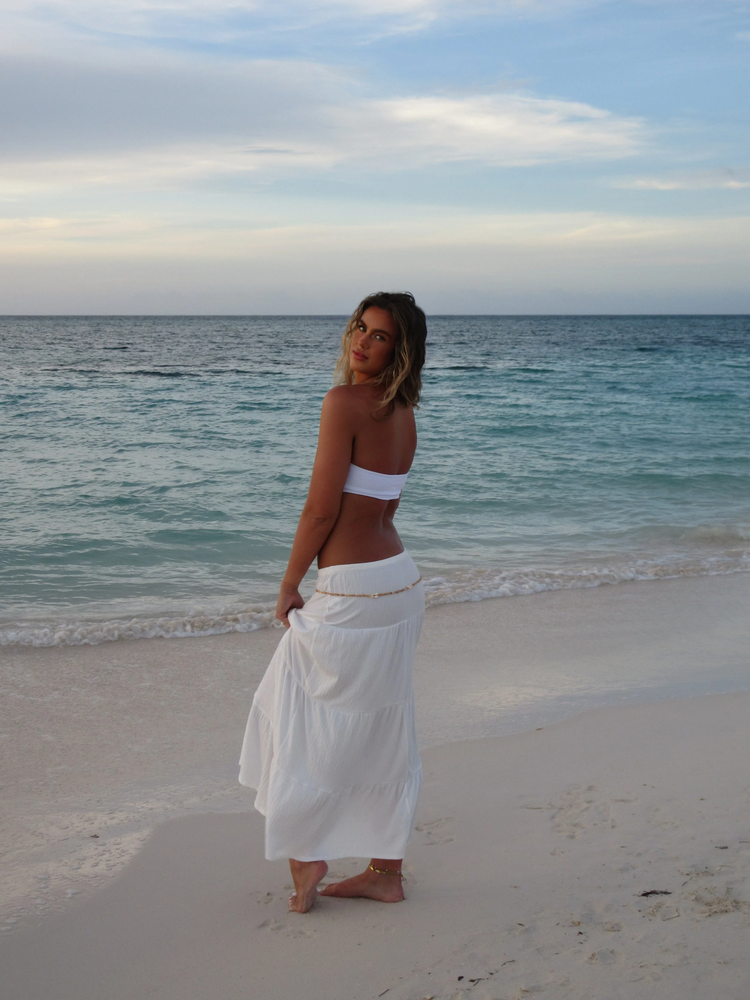 A woman in a white skirt and bandeau top standing barefoot on the beach by the ocean with a cloudy sky in the background.