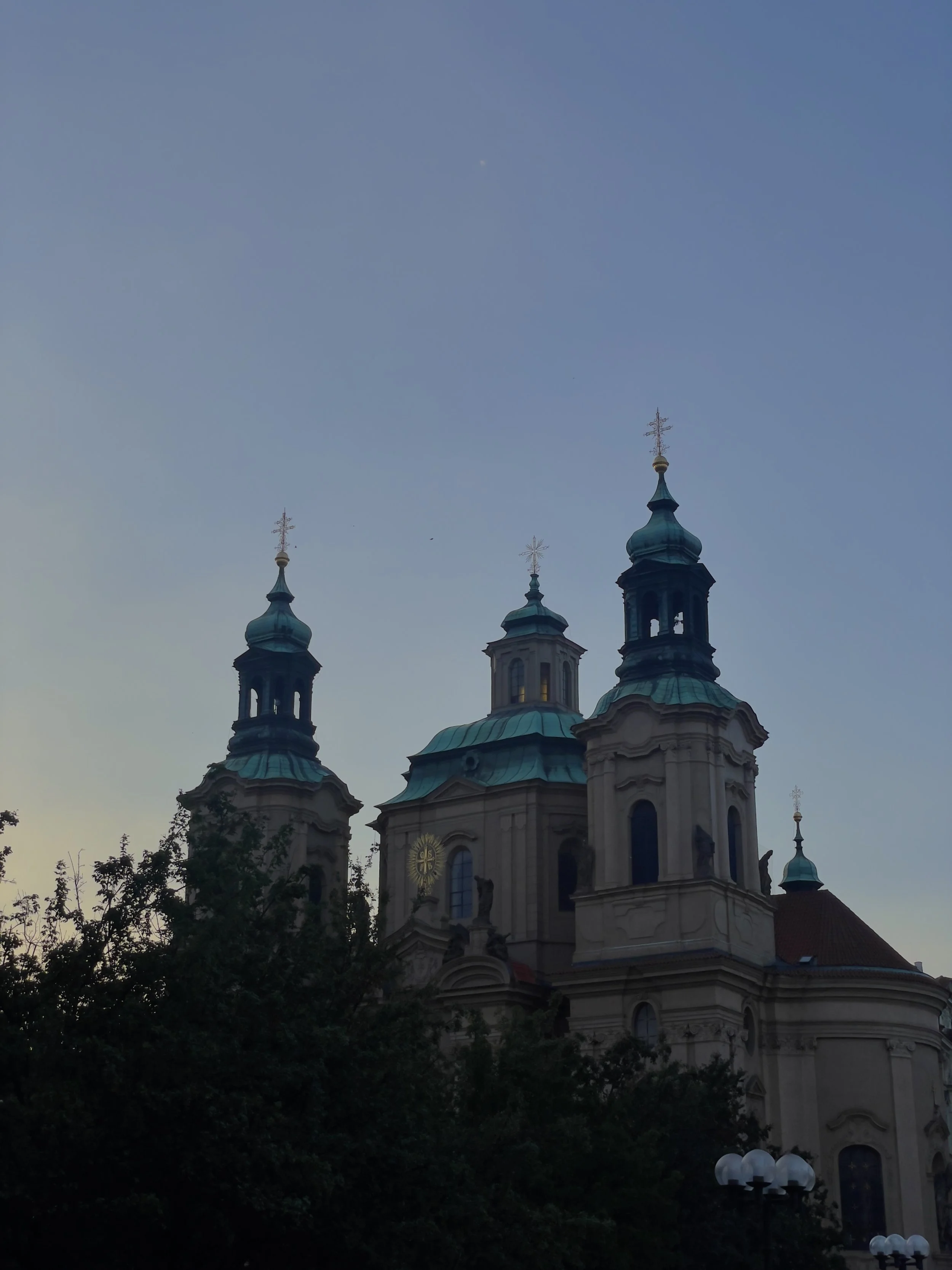 Baroque-style church with three blue domes and gold crosses on top, silhouetted against a clear evening sky, partly obscured by dark trees in the foreground.