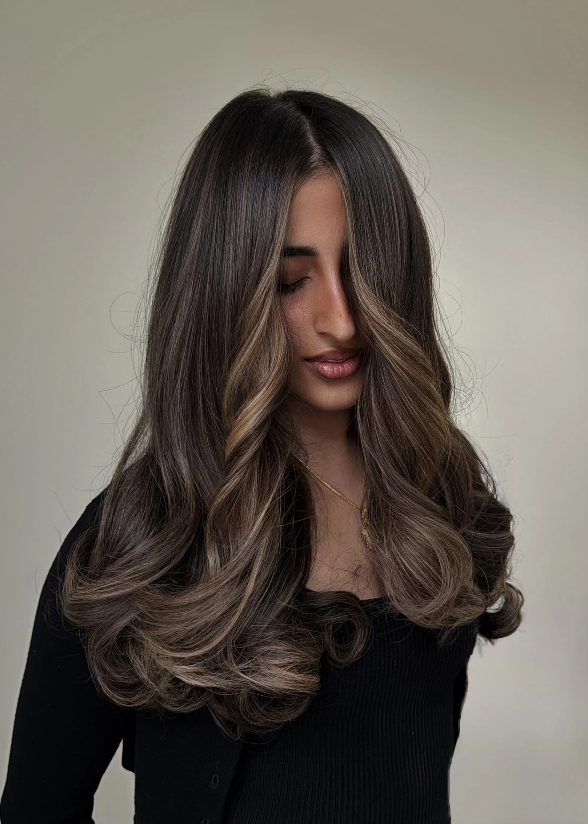 A woman with long, wavy brown hair with lighter highlights, looking downward with eyes closed, wearing a black top and a delicate gold necklace, standing against a light-colored background.