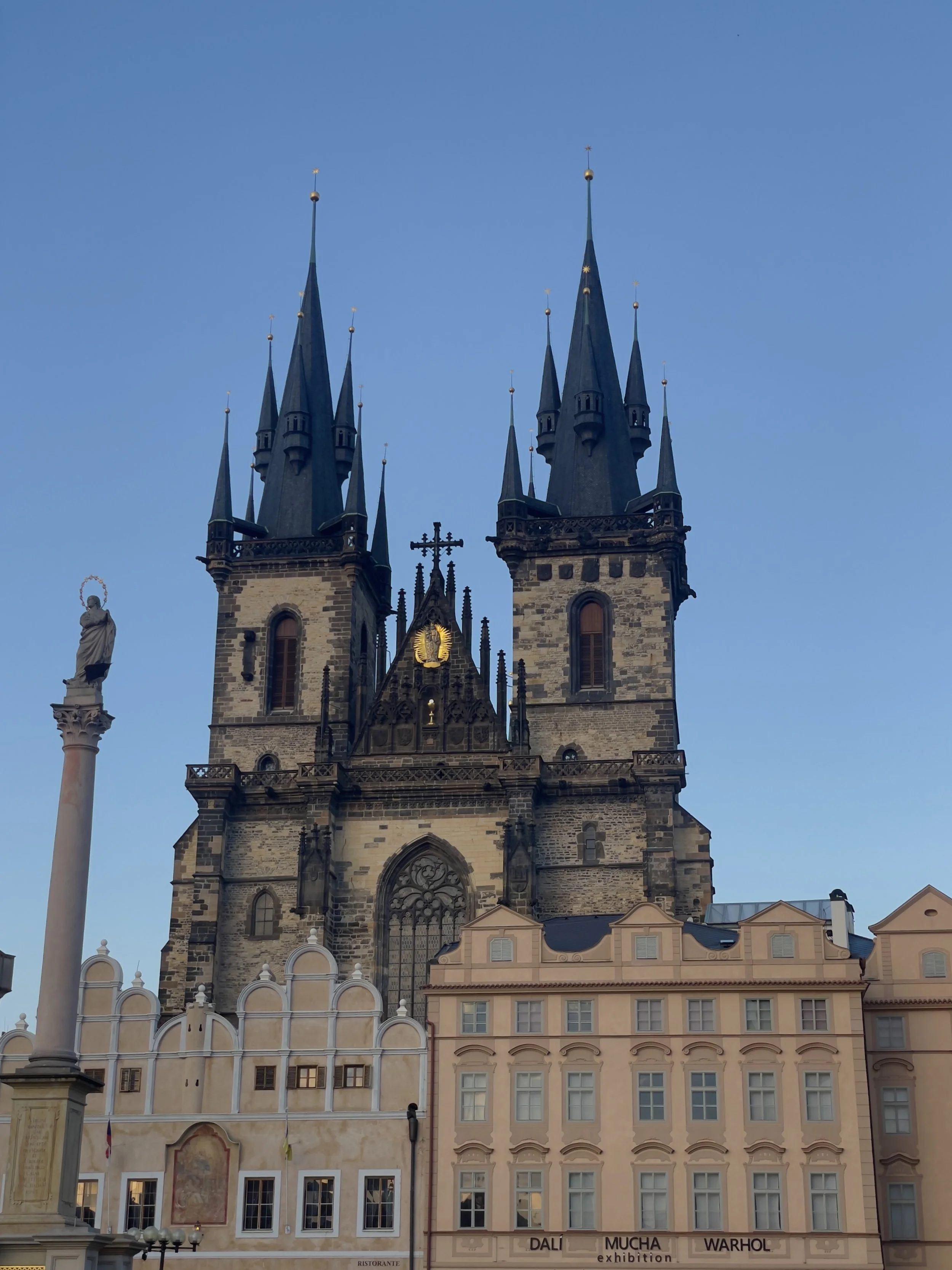A large Gothic-style church with two tall spires against a clear blue sky. There is a statue on a tall column in front of the church and pastel-colored buildings at the base.