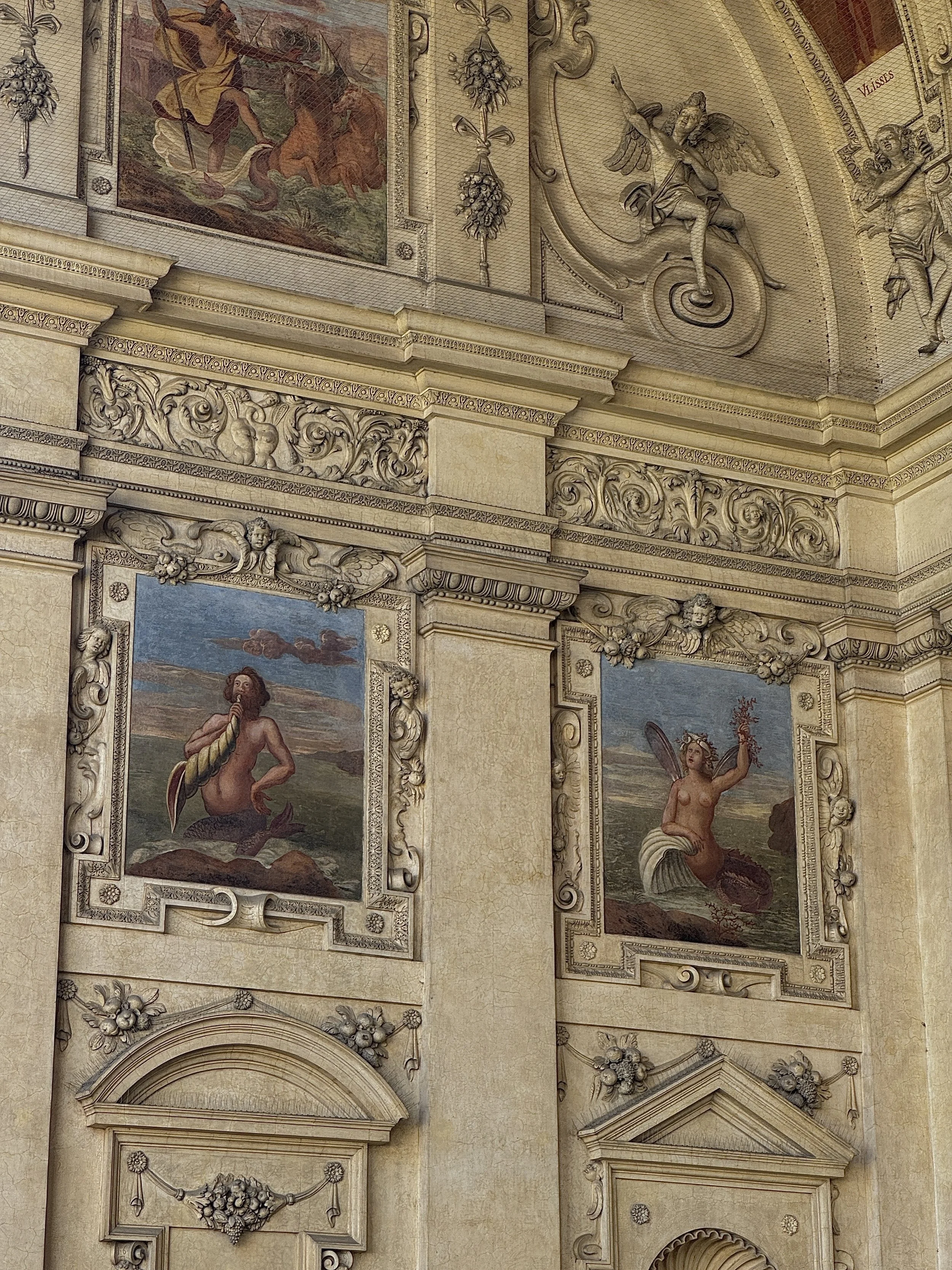 Ornate interior wall with classical murals and intricate decorative carvings. Two painted panels depict mythological water nymphs or mermaids, one holding a shell and the other holding a branch with flowers, surrounded by floral and scroll motifs.