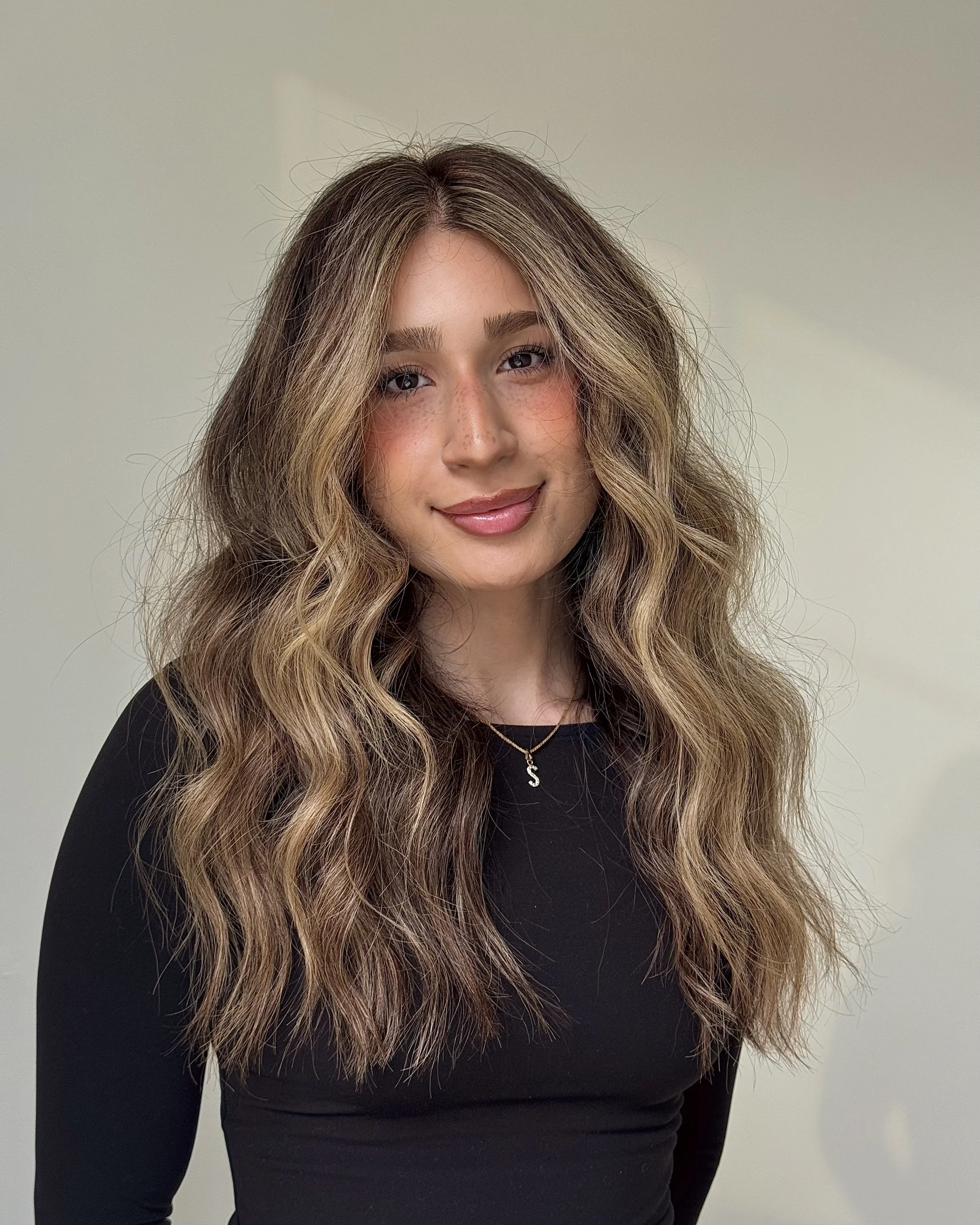 Young woman with long wavy hair, smiling, wearing a black top and a necklace with a letter 'S' pendant.
