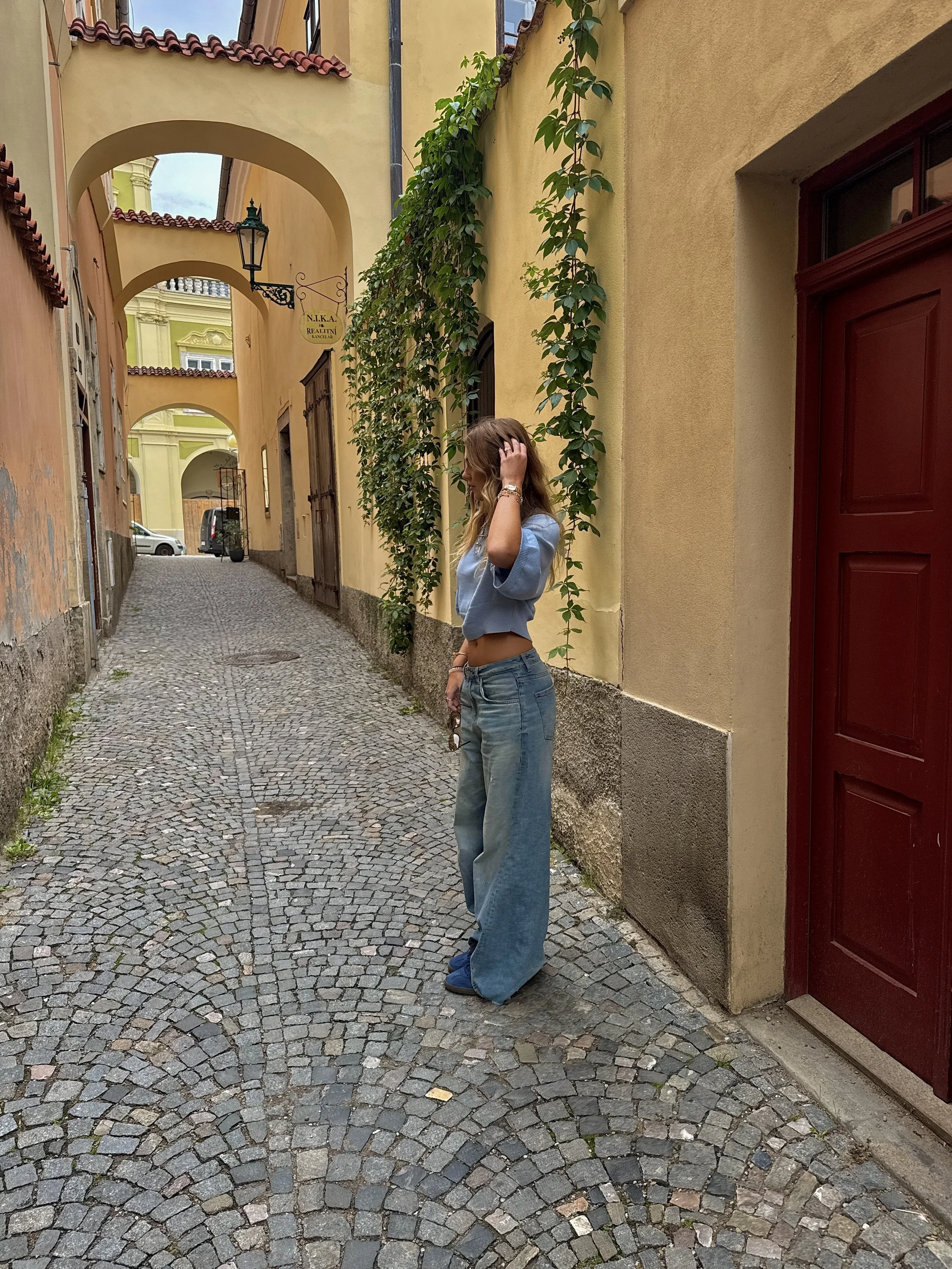 A young woman with light brown hair, wearing a light blue crop top, wide-legged jeans, and blue shoes, is standing on a cobblestone street next to a yellow building with a red door. She is touching her hair with her right hand and holding sunglasses in her left hand. There are green vines growing on the building wall, and there are archways and a parked vehicle in the background.