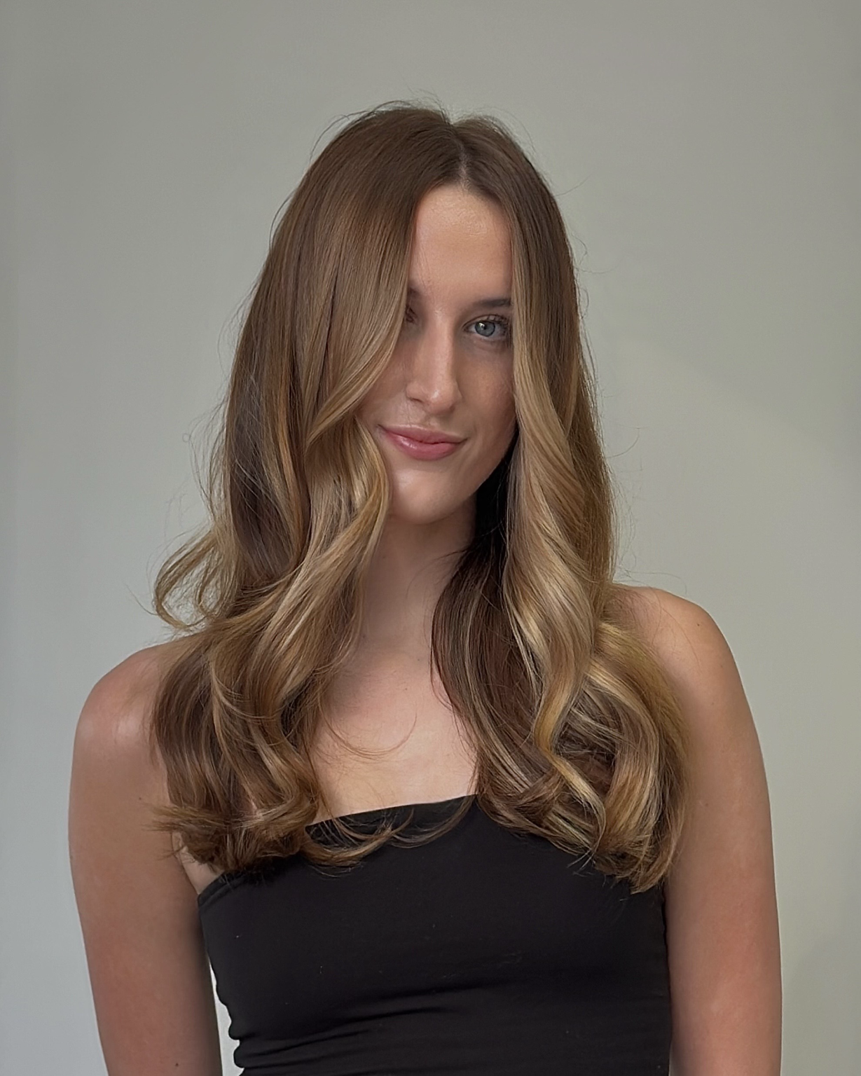 A woman with long, wavy, light brown hair and blue eyes, wearing a black strapless top, standing against a plain light-colored background.