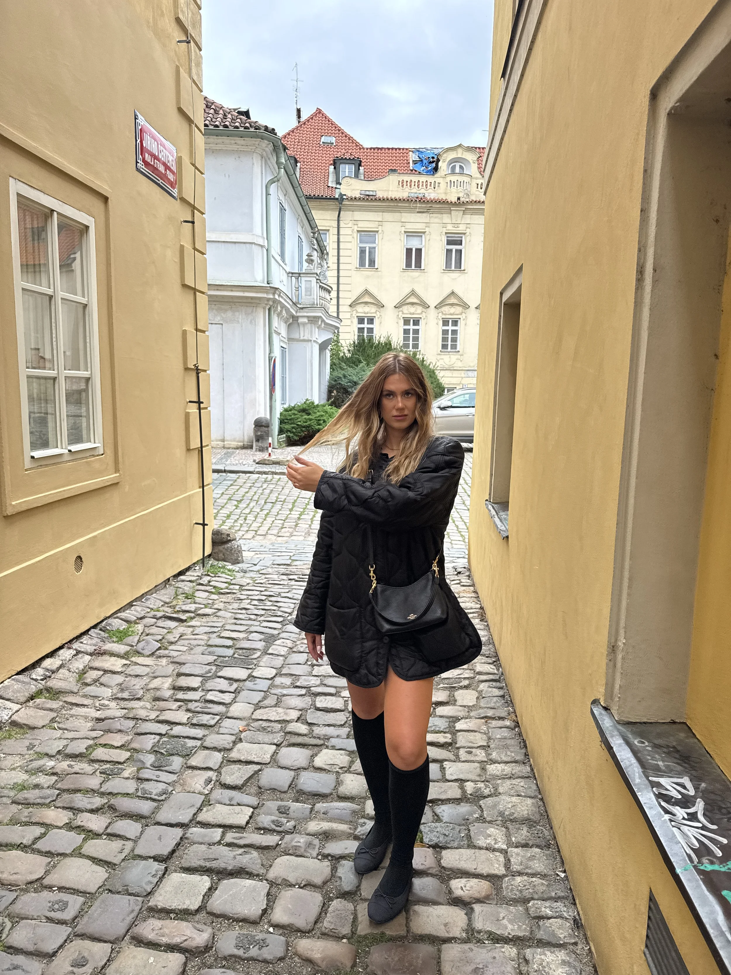A woman with long blonde hair standing on a cobblestone street between two yellow buildings, wearing a black jacket, thigh-high socks, and black shoes, with a black shoulder bag.