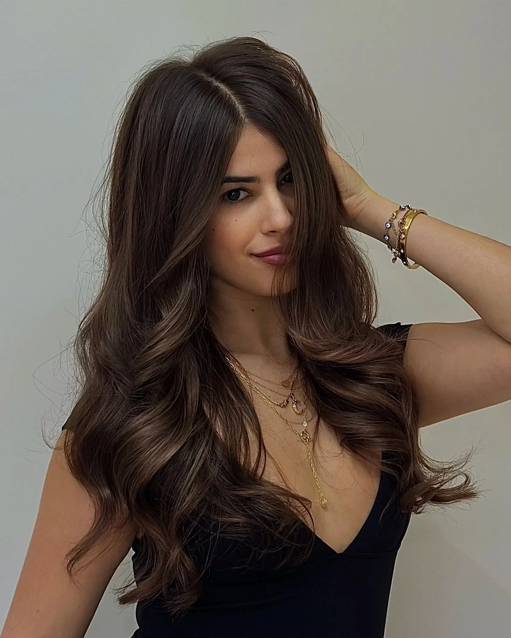 A young woman with long, wavy brown hair, wearing a black top and layered gold necklaces, with bracelets on her wrist, poses with her hand behind her head against a plain background.