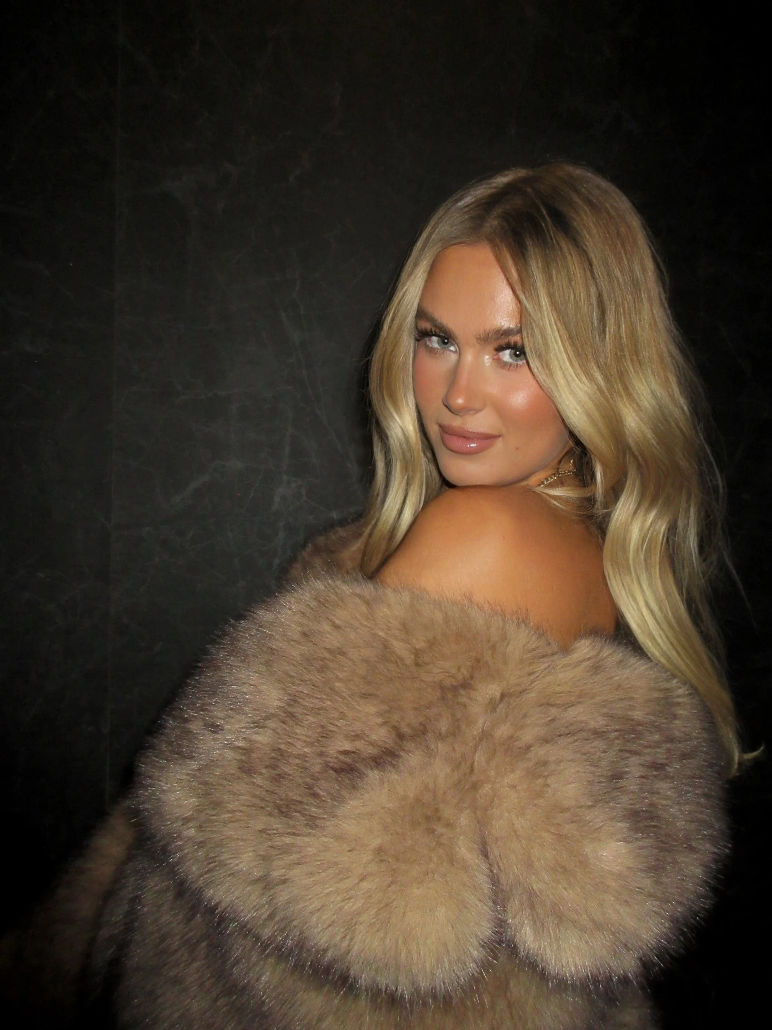 A woman with long blonde hair looking over her shoulder, draped in a fur coat, against a dark background.