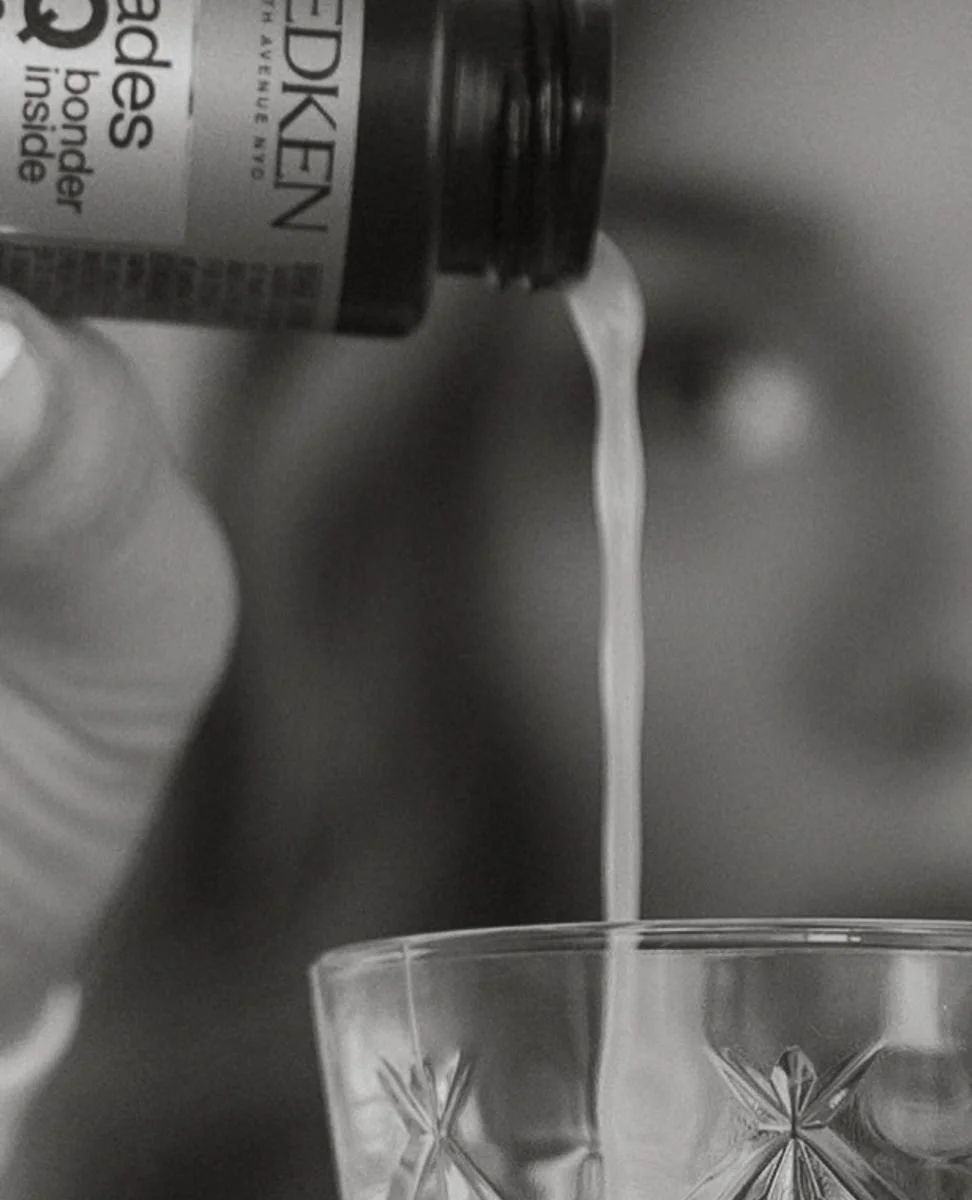 A close-up of a hand pouring a white liquid supplement from a bottle into a decorative glass.