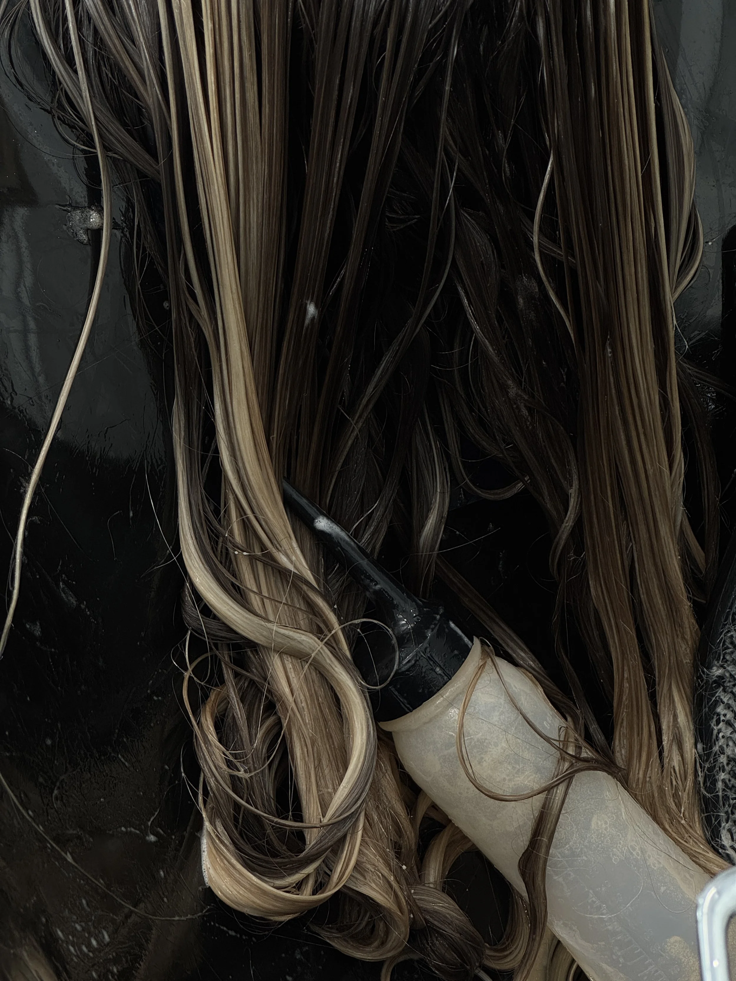 Wet, brown and blonde hair strands on a black surface with a white applicator bottle.