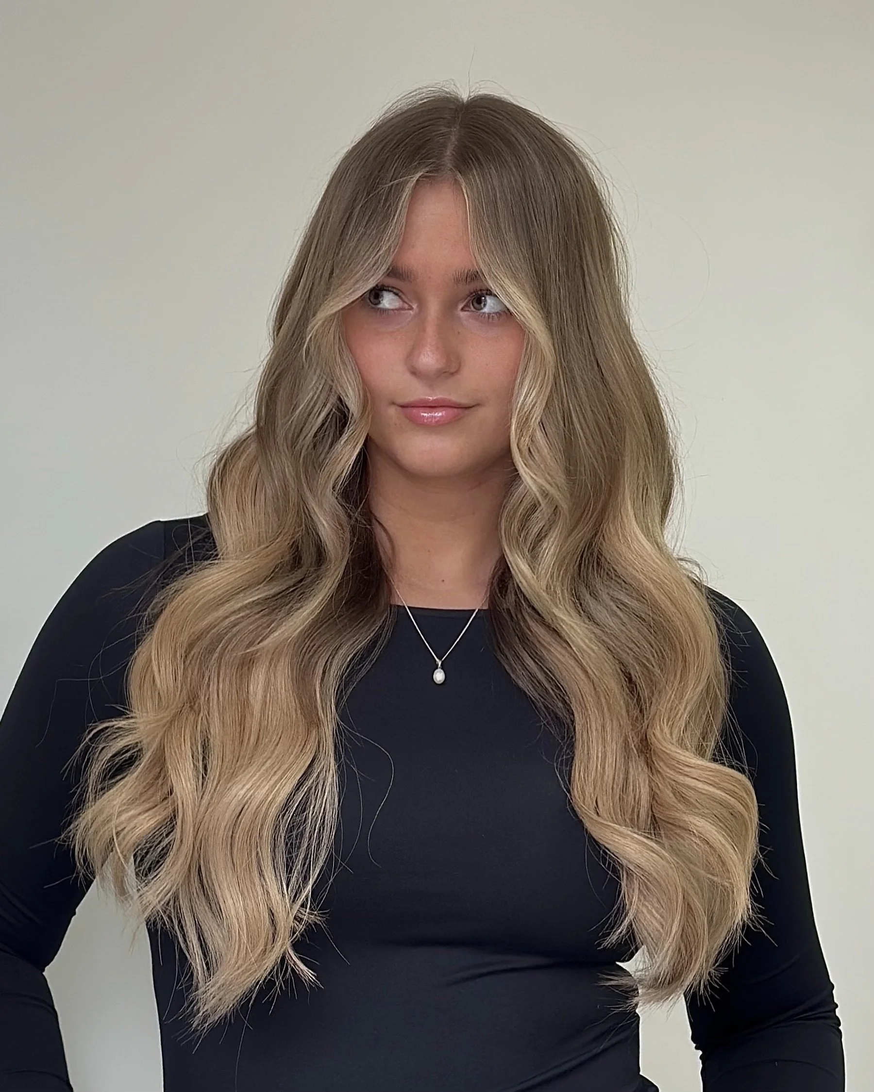 Young woman with long, wavy blonde hair wearing a black top and a silver necklace, looking to the side against a plain light-colored background.
