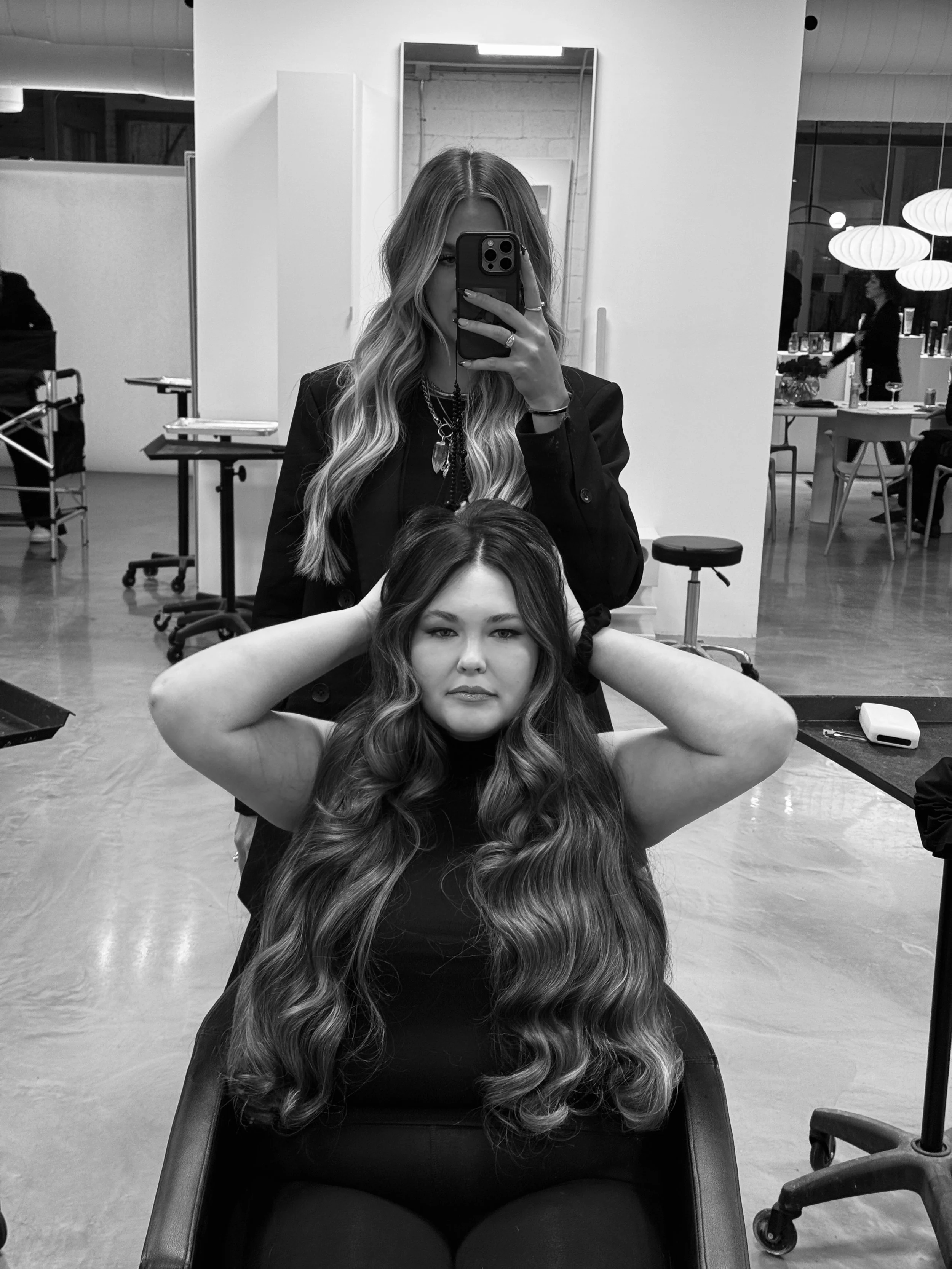 A woman with long wavy hair sits in a salon chair with her hands behind her head, while another woman with long wavy hair stands behind her, taking a selfie in a mirror.