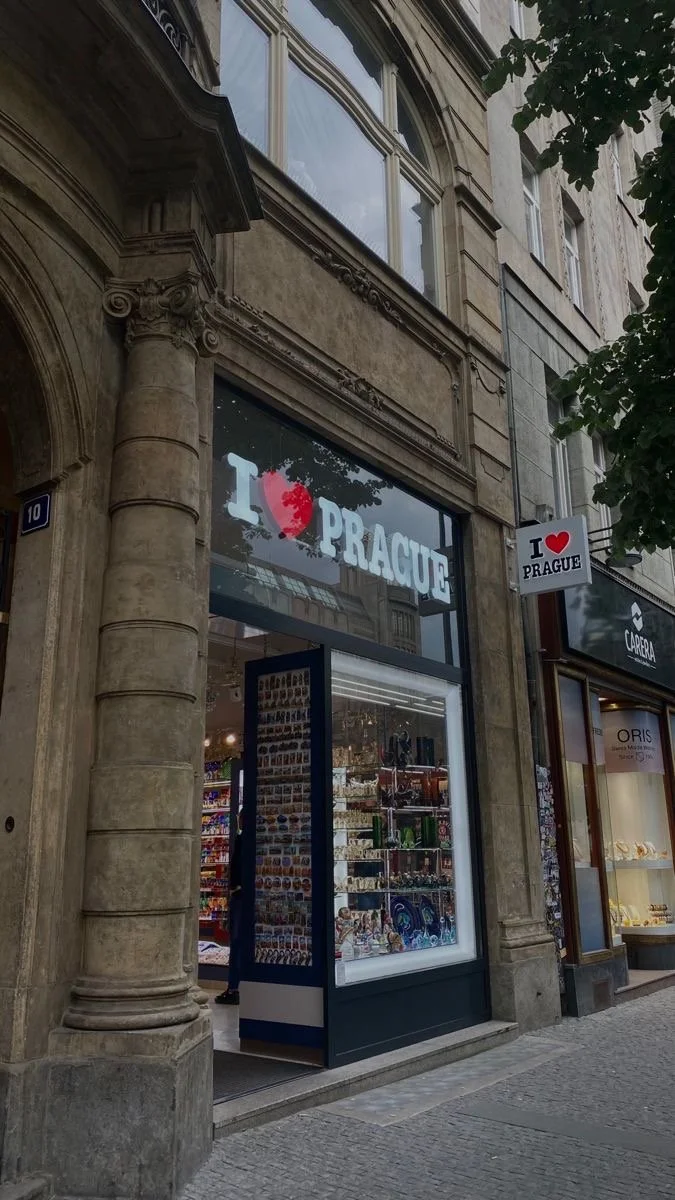 Storefront with a sign that reads "I ♥ PRAGUE" and a large display window showing souvenirs inside. Next door is a jewelry store named ORIS.