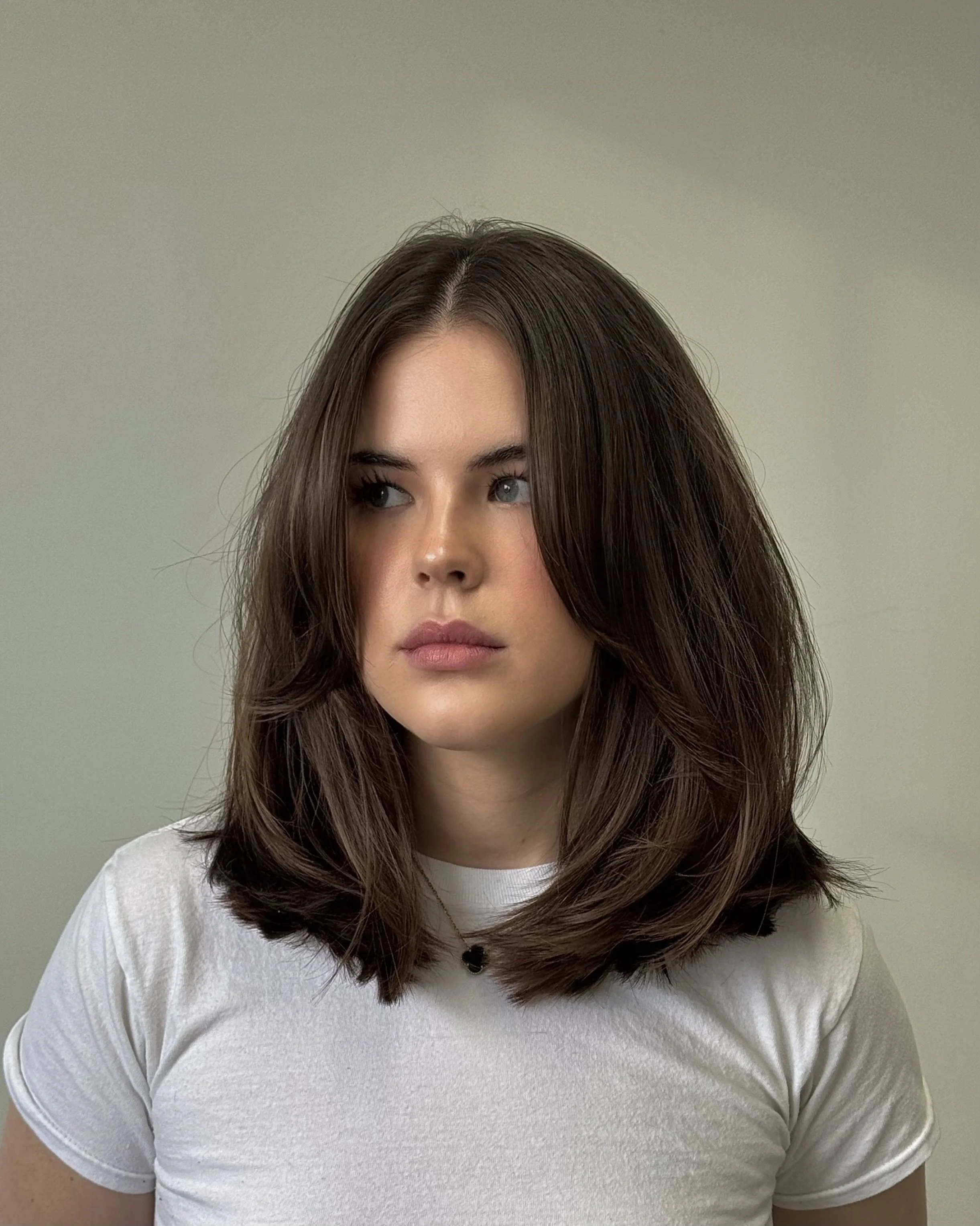 A woman with shoulder-length brown hair styled in loose waves, wearing a white t-shirt and a small black necklace, looking off to the side with a neutral expression against a plain gray background.