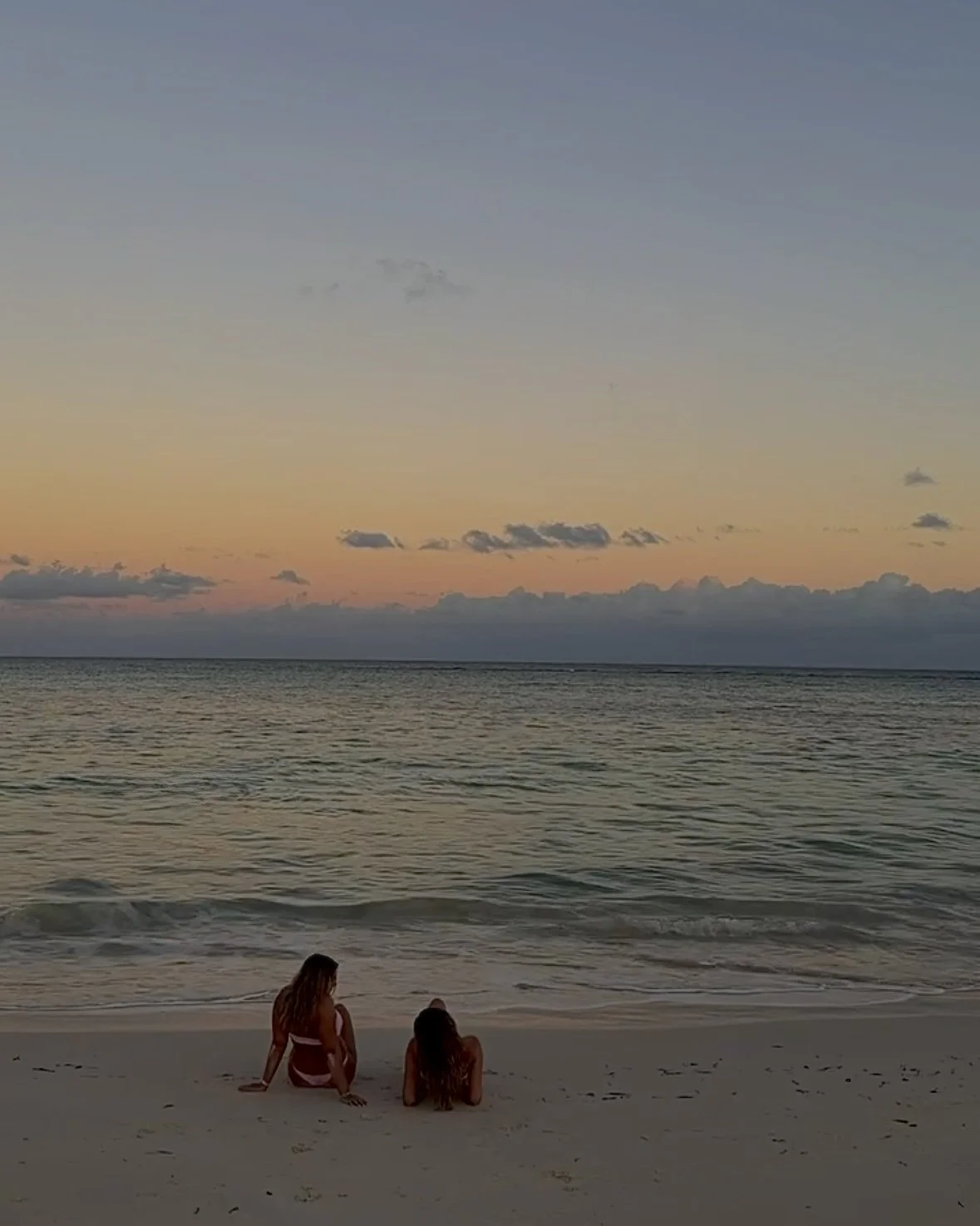 Two women sitting on a sandy beach at sunset, looking at the ocean, with a sky featuring soft pastel colors and a few clouds.