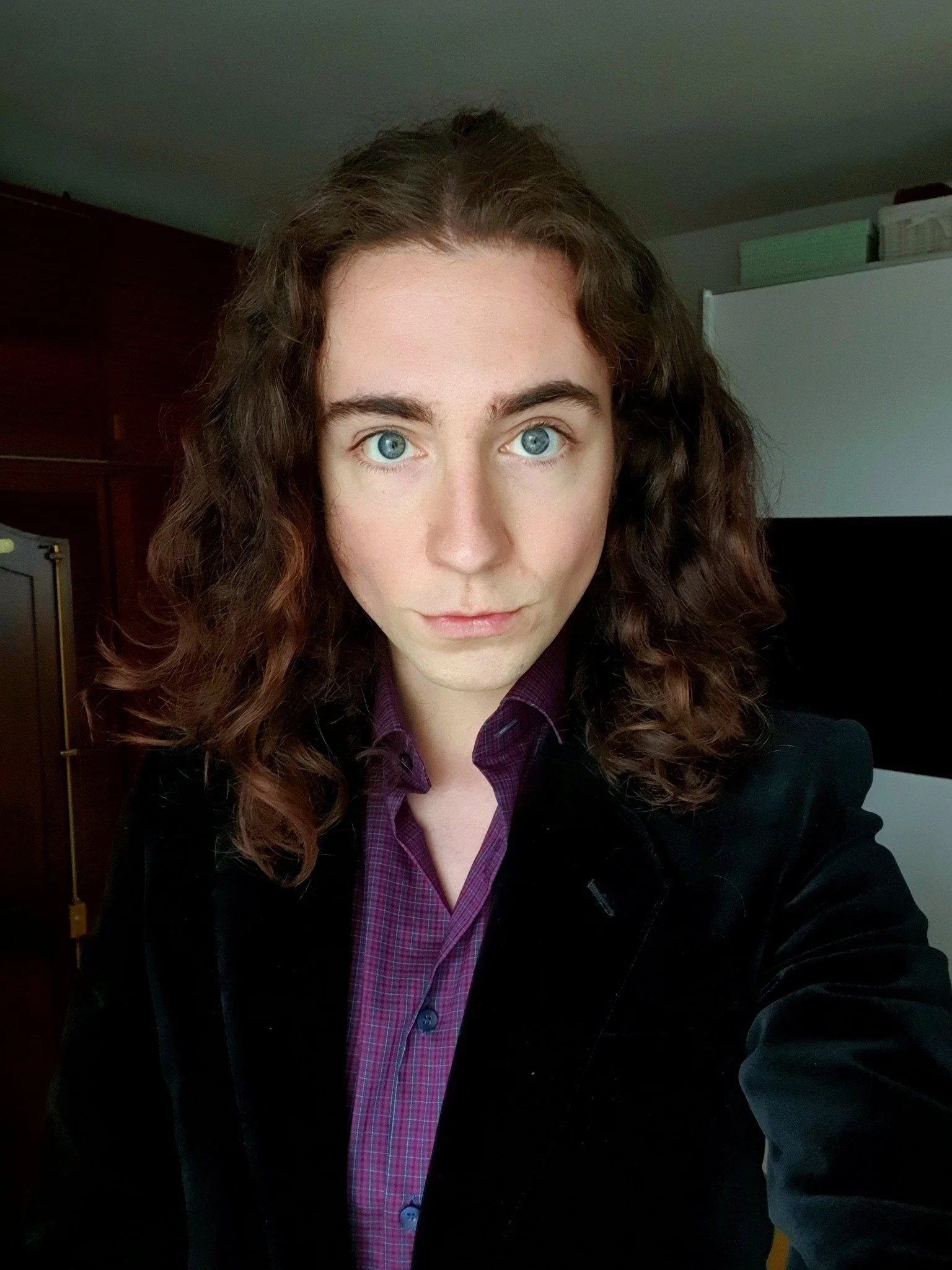 Close-up of a woman with curly brown hair and blue eyes, wearing a dark blazer over a purple plaid shirt, indoors with a dark wooden cabinet and a white cabinet in the background.