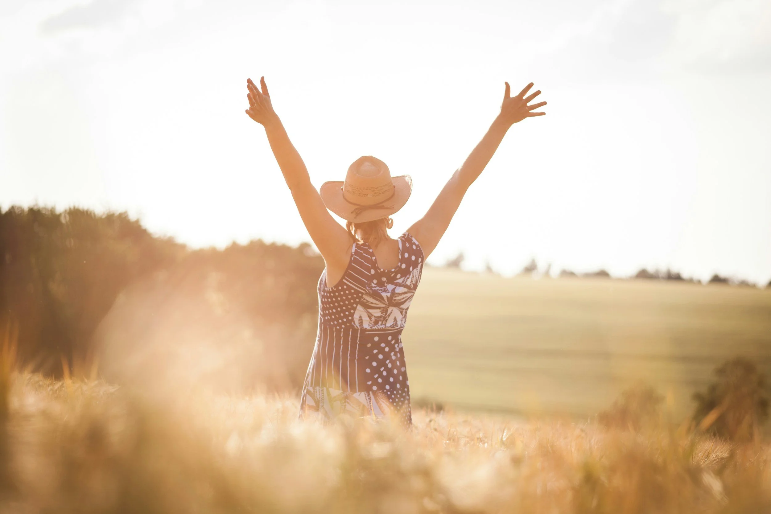 A woman in a polka dot dress and straw hat standing in a field with arms raised towards the sky, sunlight and open sky in the background