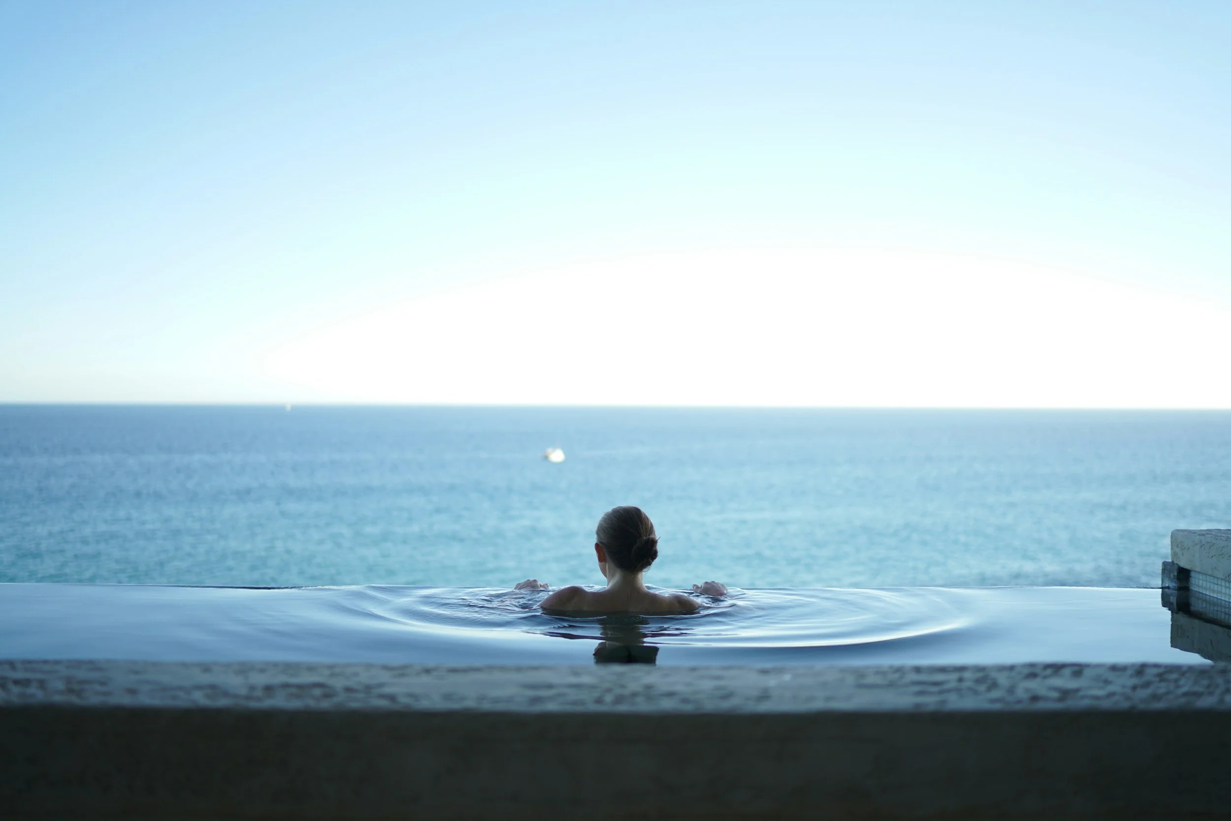 A woman with her hair tied back relaxing in an infinity pool overlooking the ocean on a sunny day.