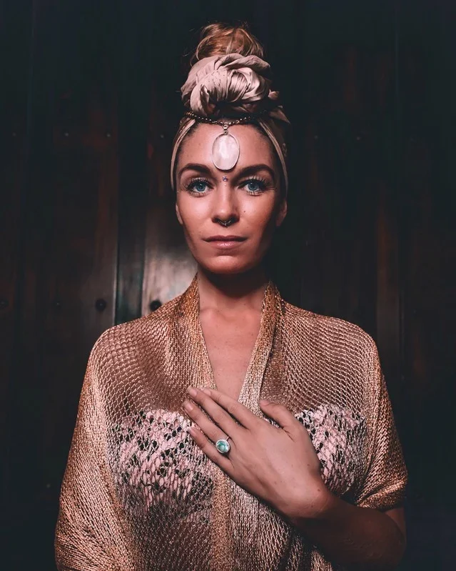 A woman with blue eyes and blonde hair wrapped in a beige headscarf adorned with a large gemstone, wearing a gold metallic shawl with pink embroidery, and a large ring with a turquoise stone on her left hand, standing in front of a dark wooden background.